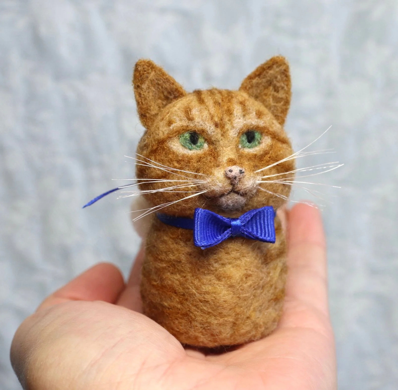 A small, realistic felted cat figure with orange fur, green eyes, whiskers, and a blue bow tie, being held in a person's hand.