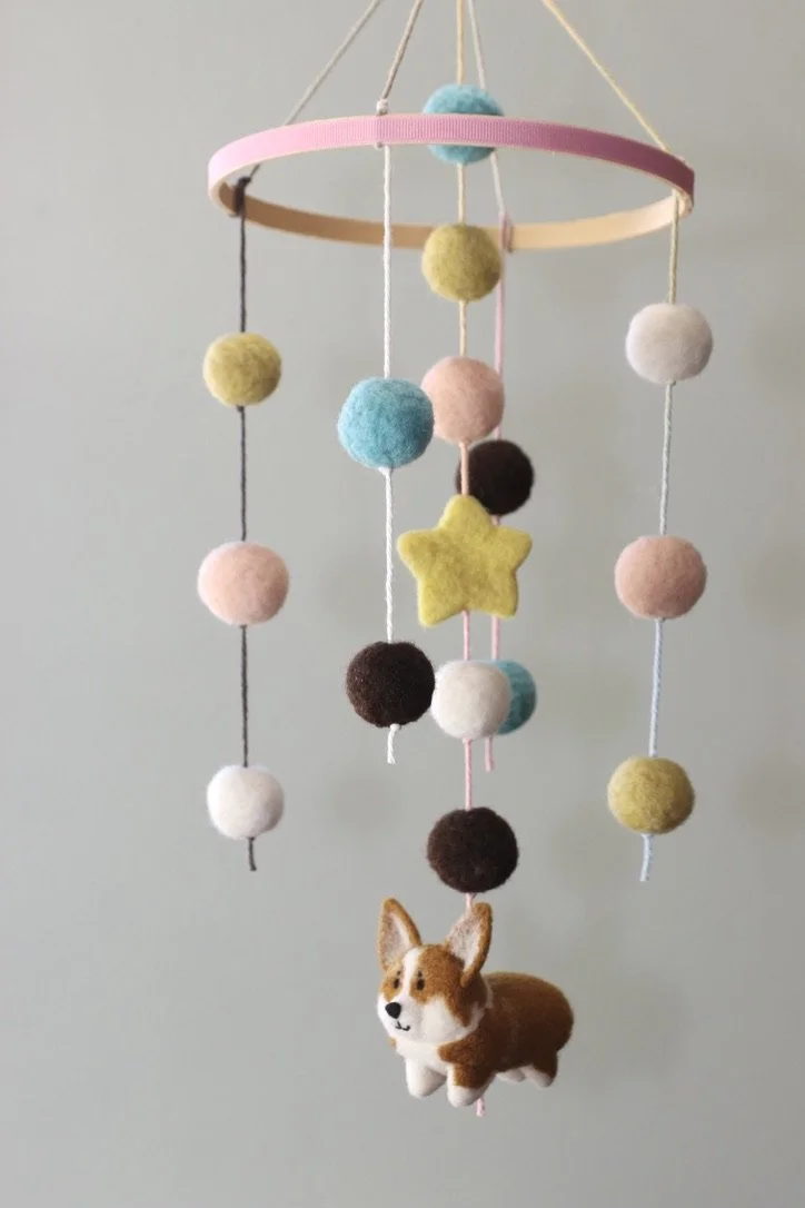 Colorful felt mobile with hanging pom-poms, a star, and a plush dog at the bottom, suspended from a pink and wooden circular frame.