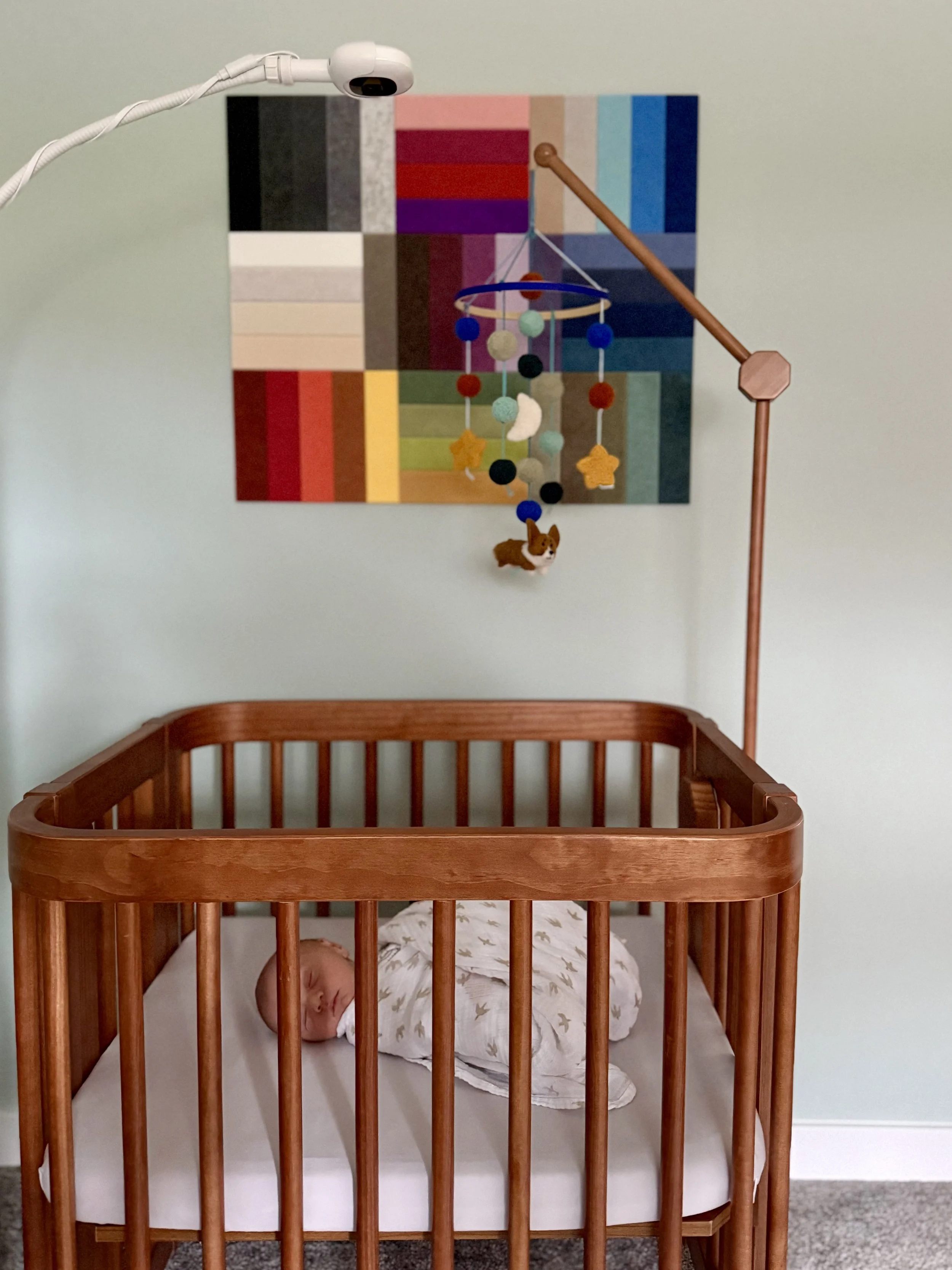 A baby sleeping in a wooden crib against a light green wall. Above, a colorful palette-themed artwork and a mobile hanging from a lamp arm.