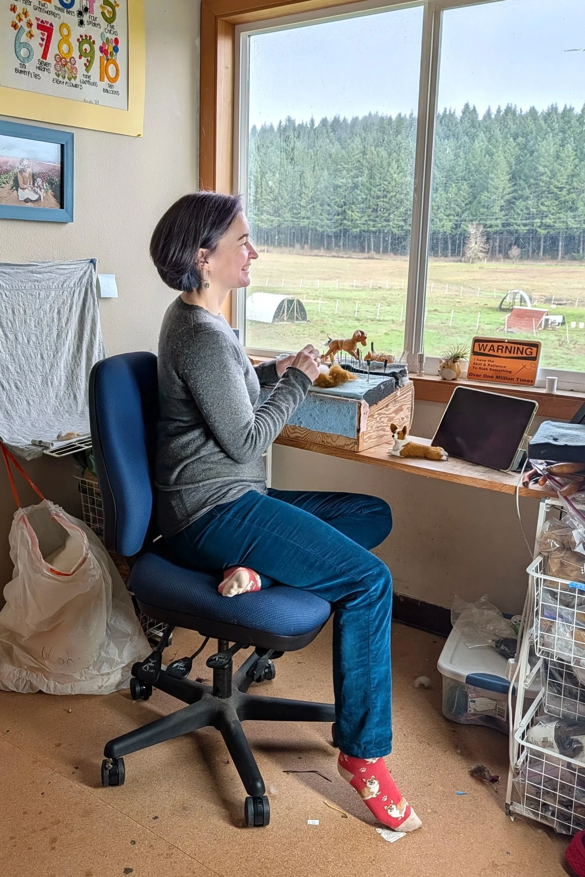 Woman sitting in office chair at desk, working on miniature dog figurines, with a large window showing a rural farm landscape with trees and pastures.