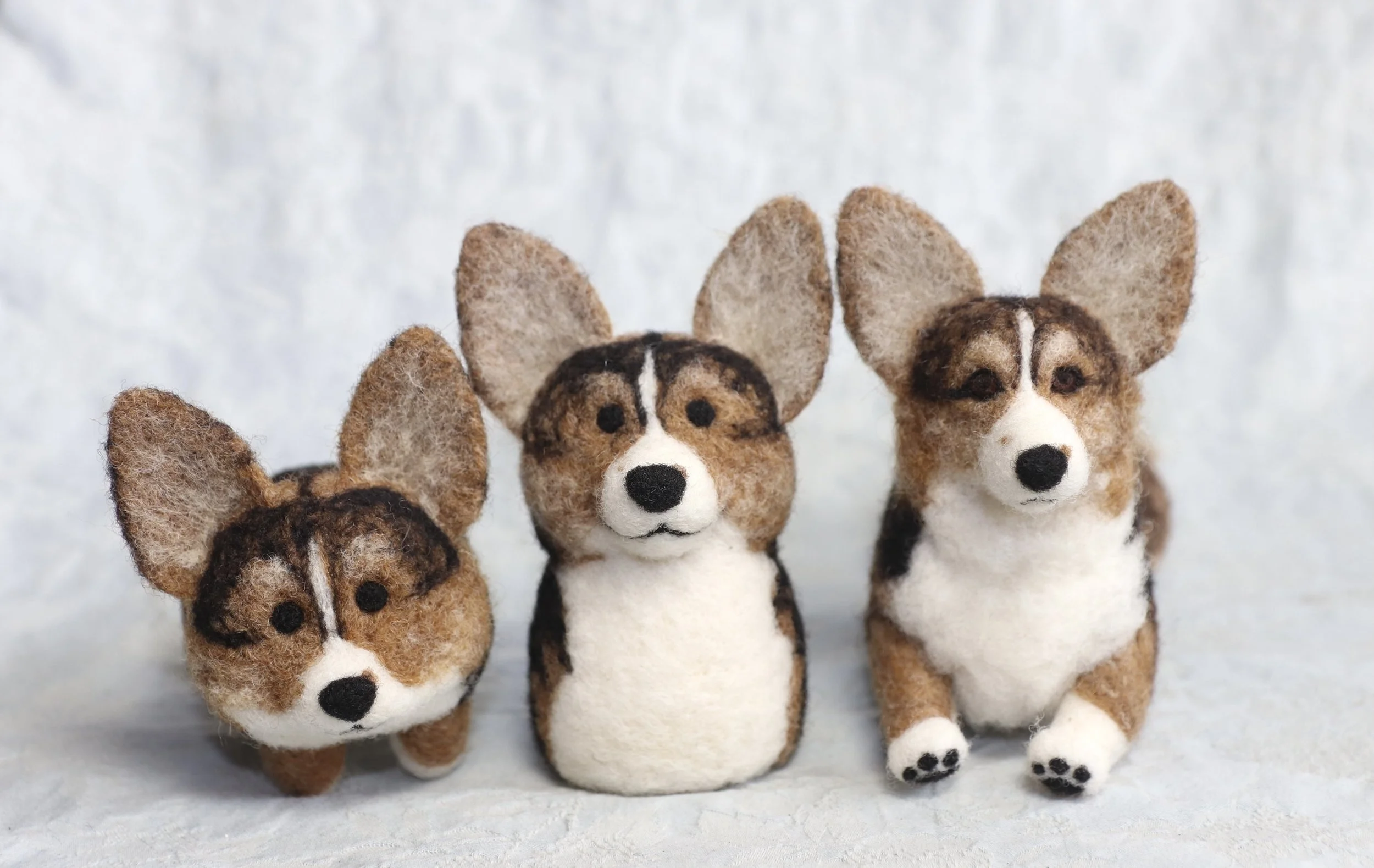 Three felted dog figurines resembling corgis with large ears, black and white facial markings, and brown, black, and white fur, set against a light background.