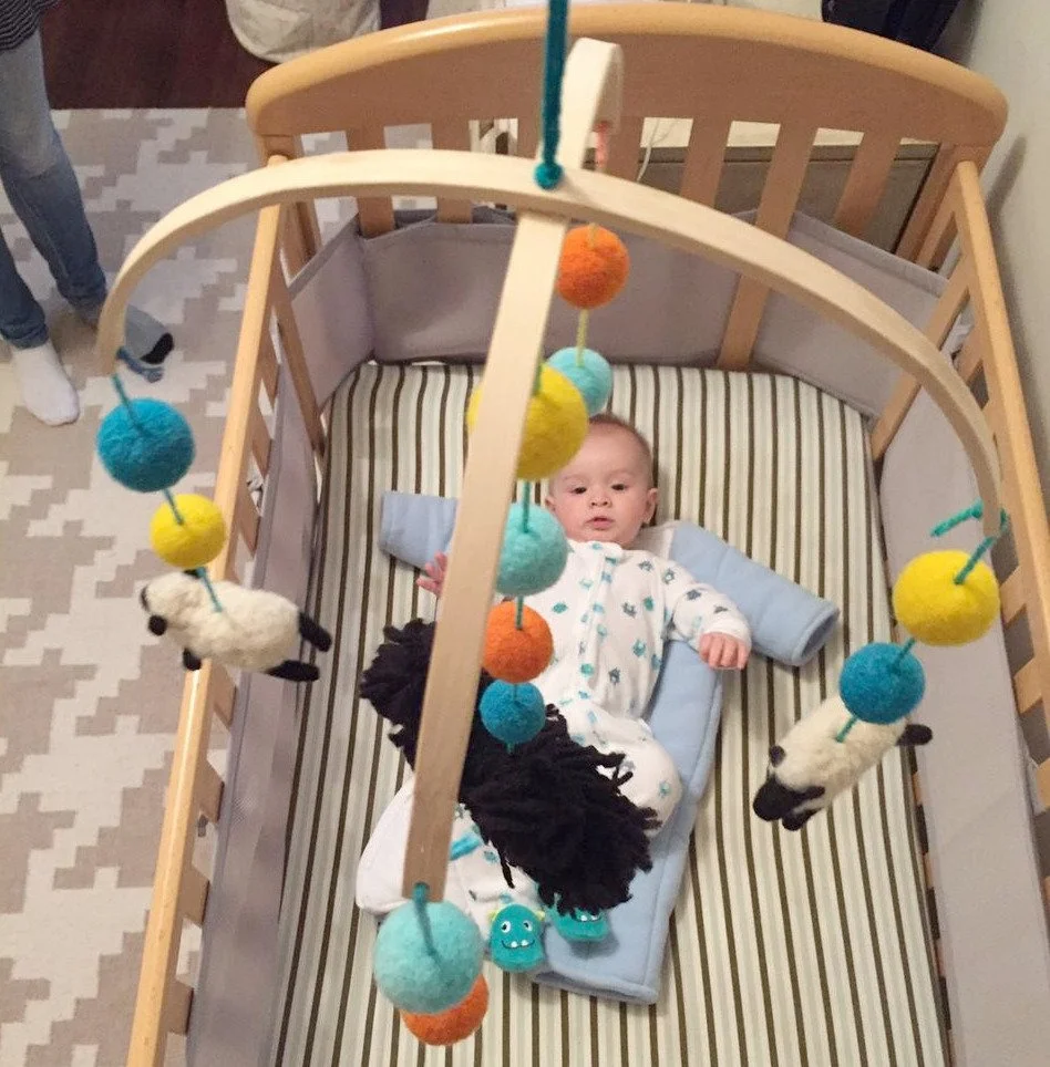 A baby lying in a crib with a striped mattress, holding a blanket, and looking up at a mobile with colorful felt balls and small panda plush toys hanging above.