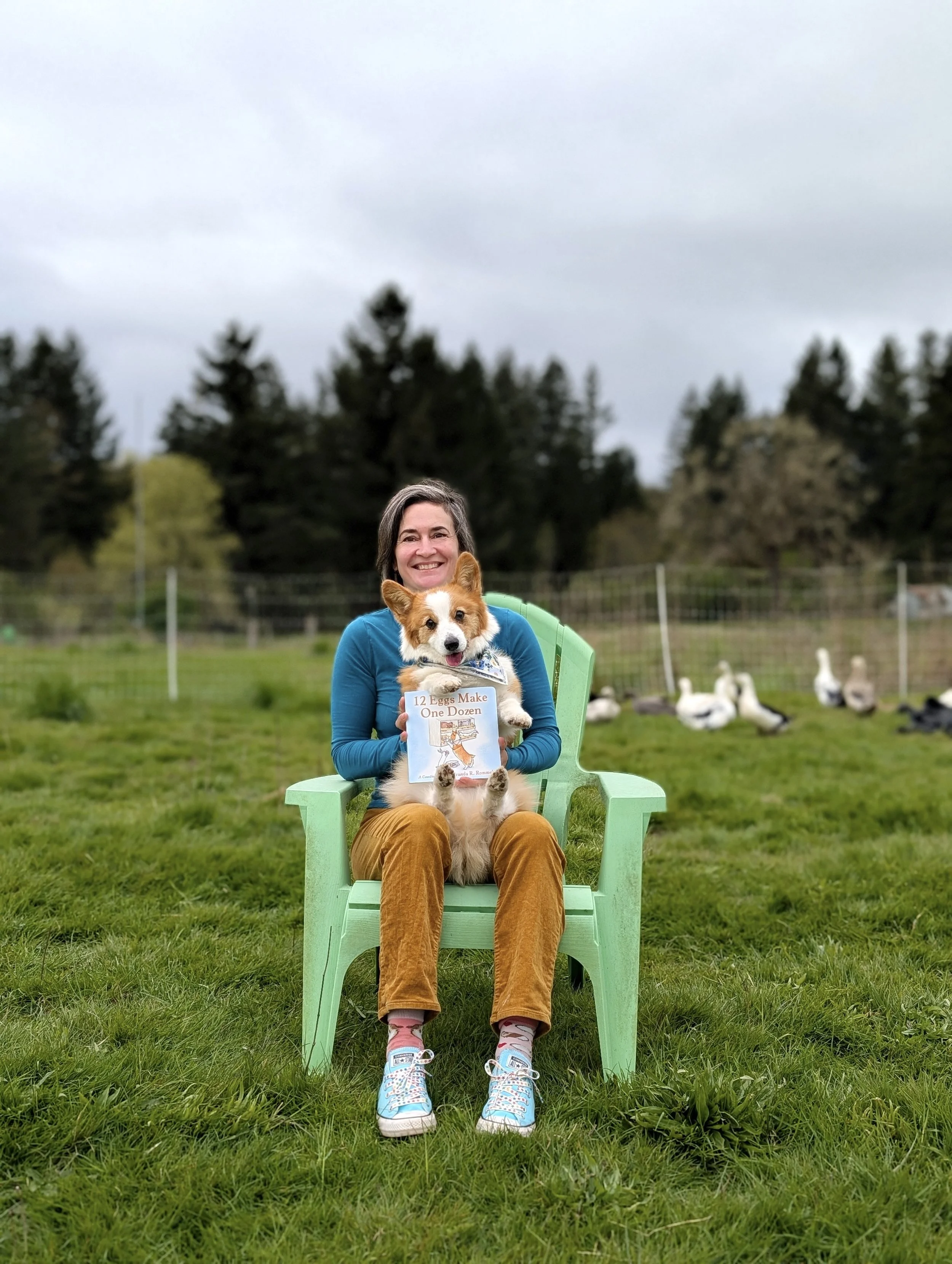 Woman sitting on a green chair outdoors, holding a small dog and a children's book, with a goose in the background.