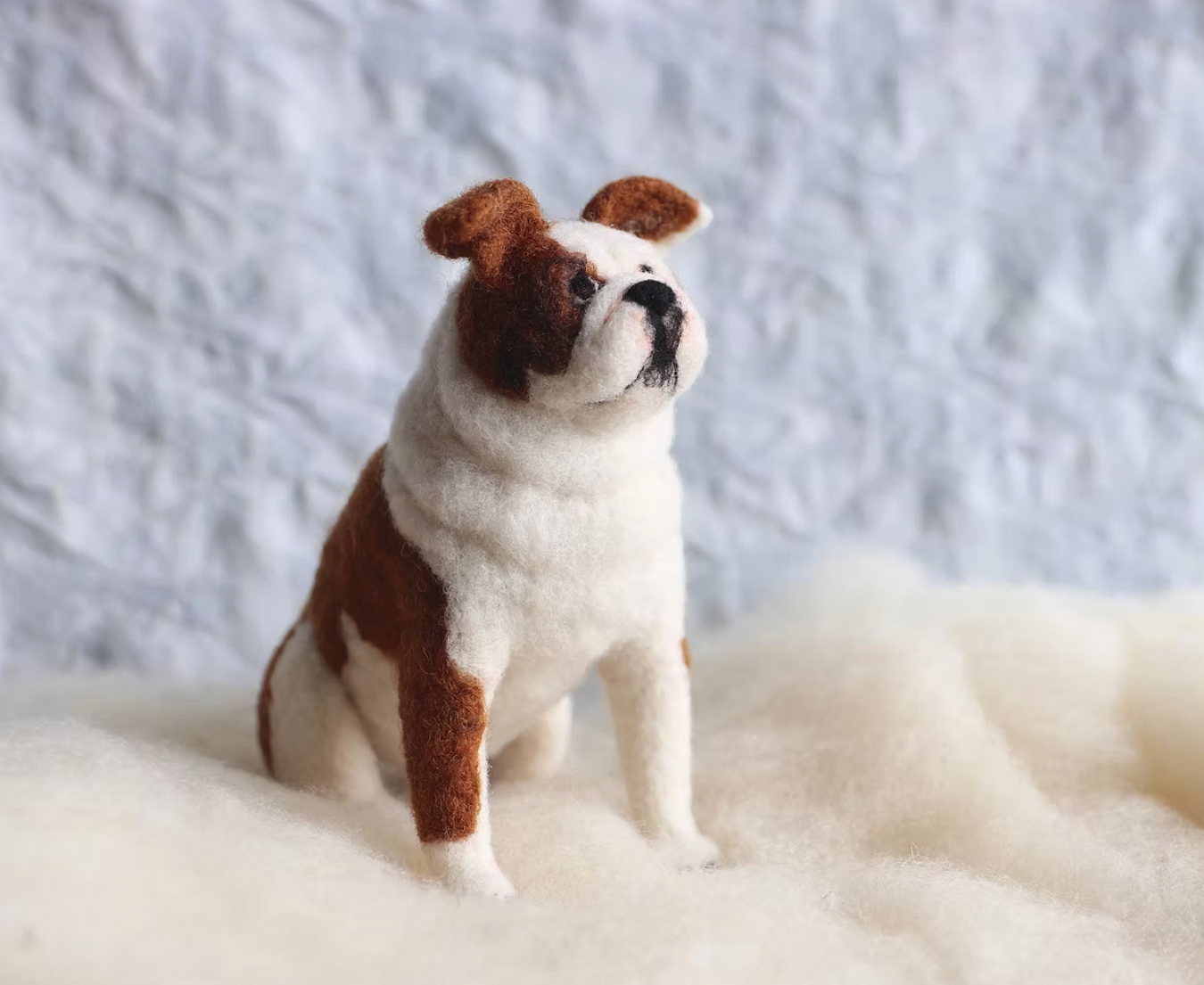 Felted dog sculpture resembling a French Bulldog sitting on white fabric with a textured white wall in the background.
