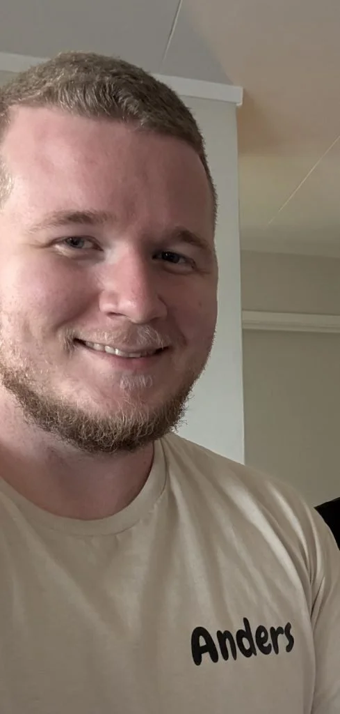 Et nærbilde av en smilende mann med kort hår, skjegg og lys hud, som har på seg en beige t-skjorte med navnet «Anders» på.
