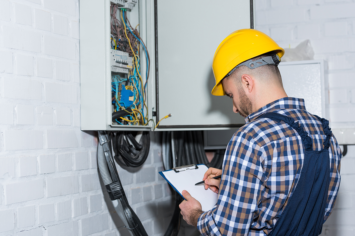 Electricity inspector wearing a yellow safety helmet and plaid shirt inspecting electrical circuit breaker panel with a clipboard and pen