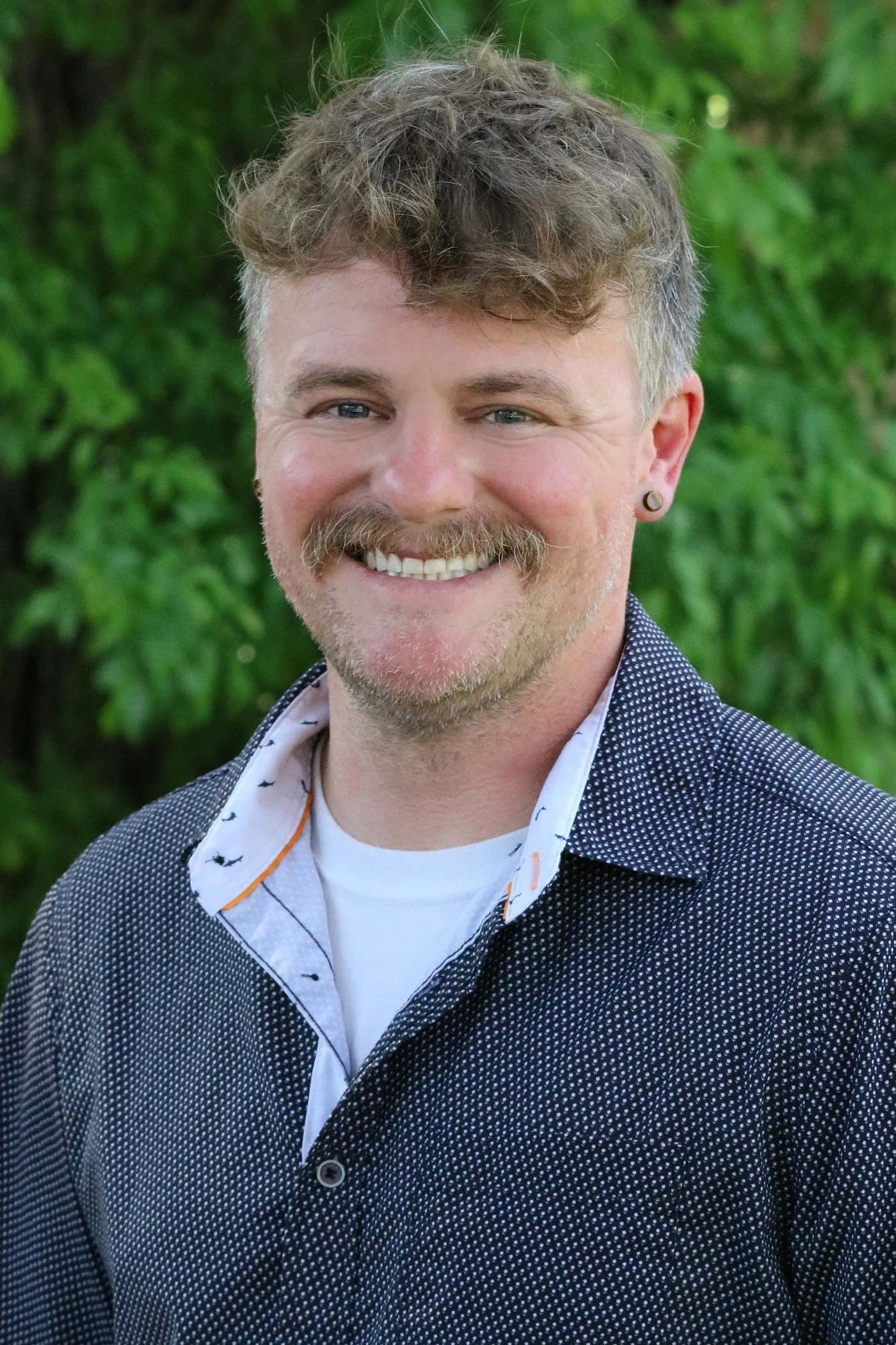 A smiling man with curly light brown hair, a mustache and beard, wearing a patterned shirt and a white undershirt, standing outdoors with green foliage in the background.