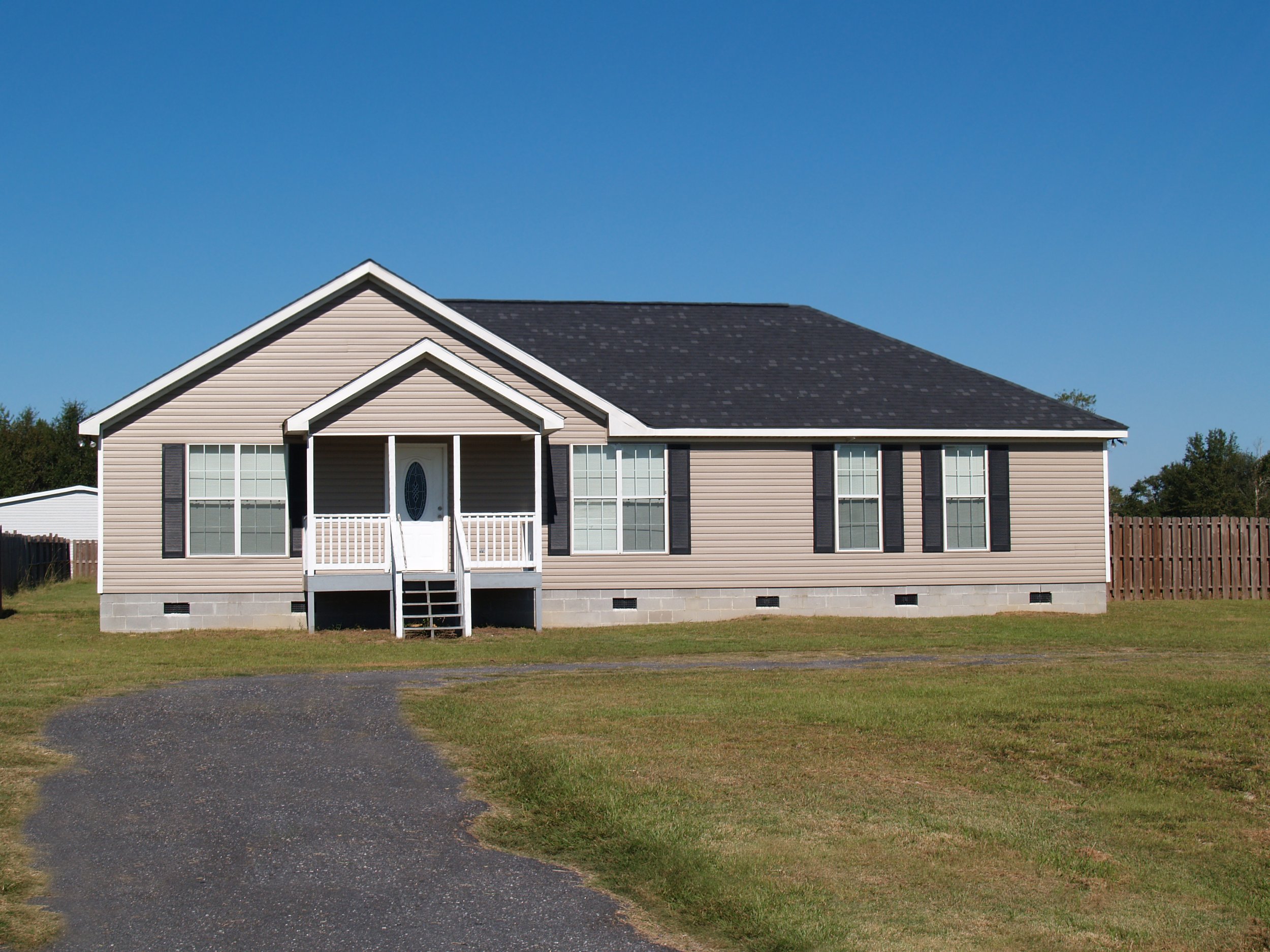 A beige single-story house with black shutters, a small covered front porch with white railing, a black shingled roof, and a curved driveway on a grassy lawn under a clear blue sky.