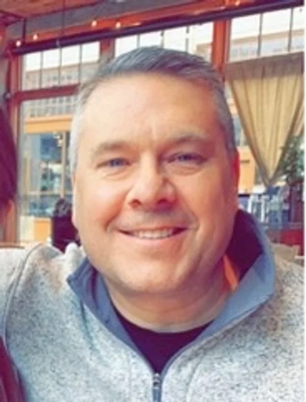 A smiling man with short black and gray hair, wearing a gray zip-up jacket, in a restaurant or cafe with red framing and large windows in the background.