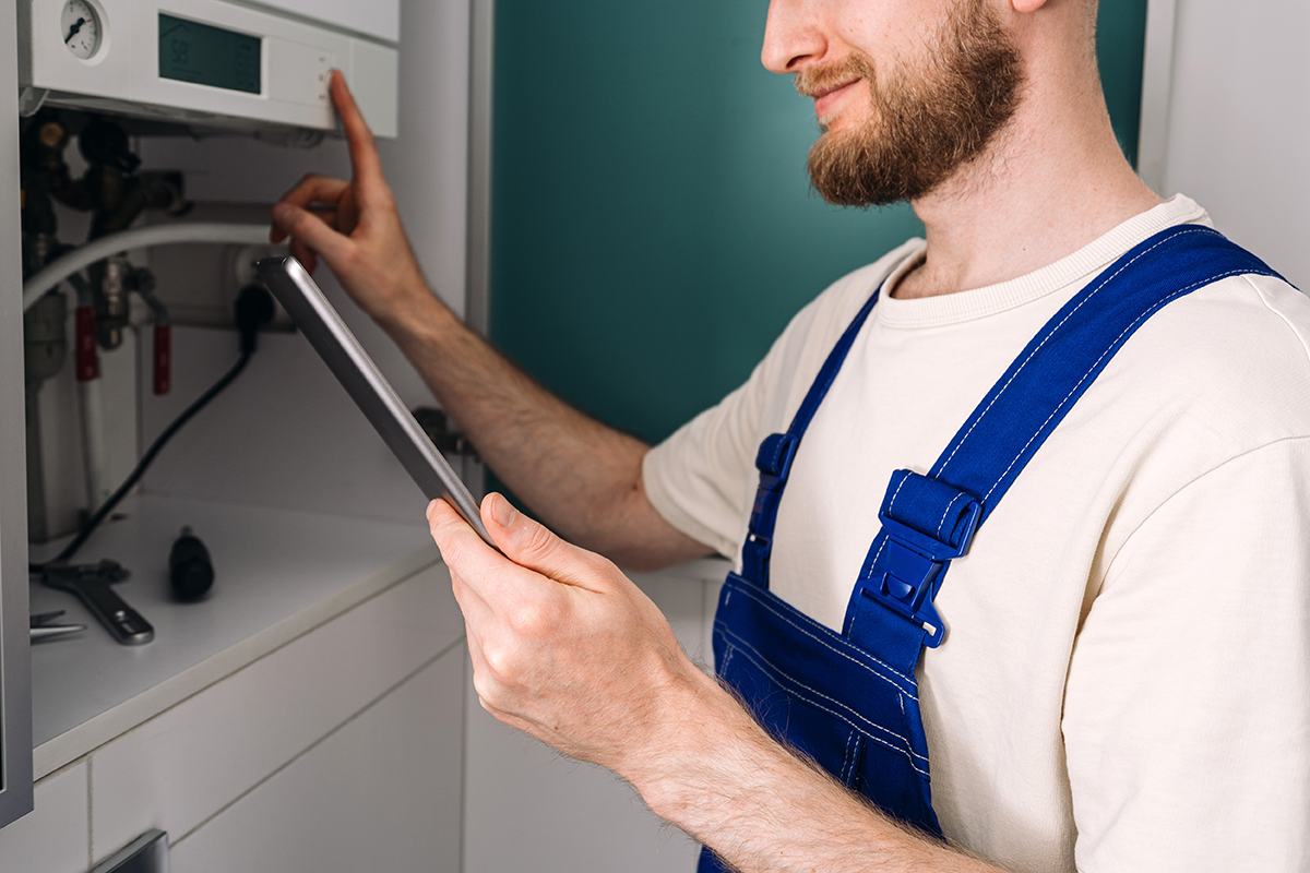 Inspector in a white shirt and blue overalls using a tablet while looking at a control panel in a utility or maintenance closet.