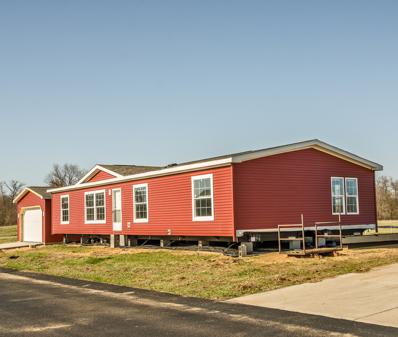 Red manufactured home with white trim on foundation blocks, set on a grassy lot with clear blue sky and trees in the background.