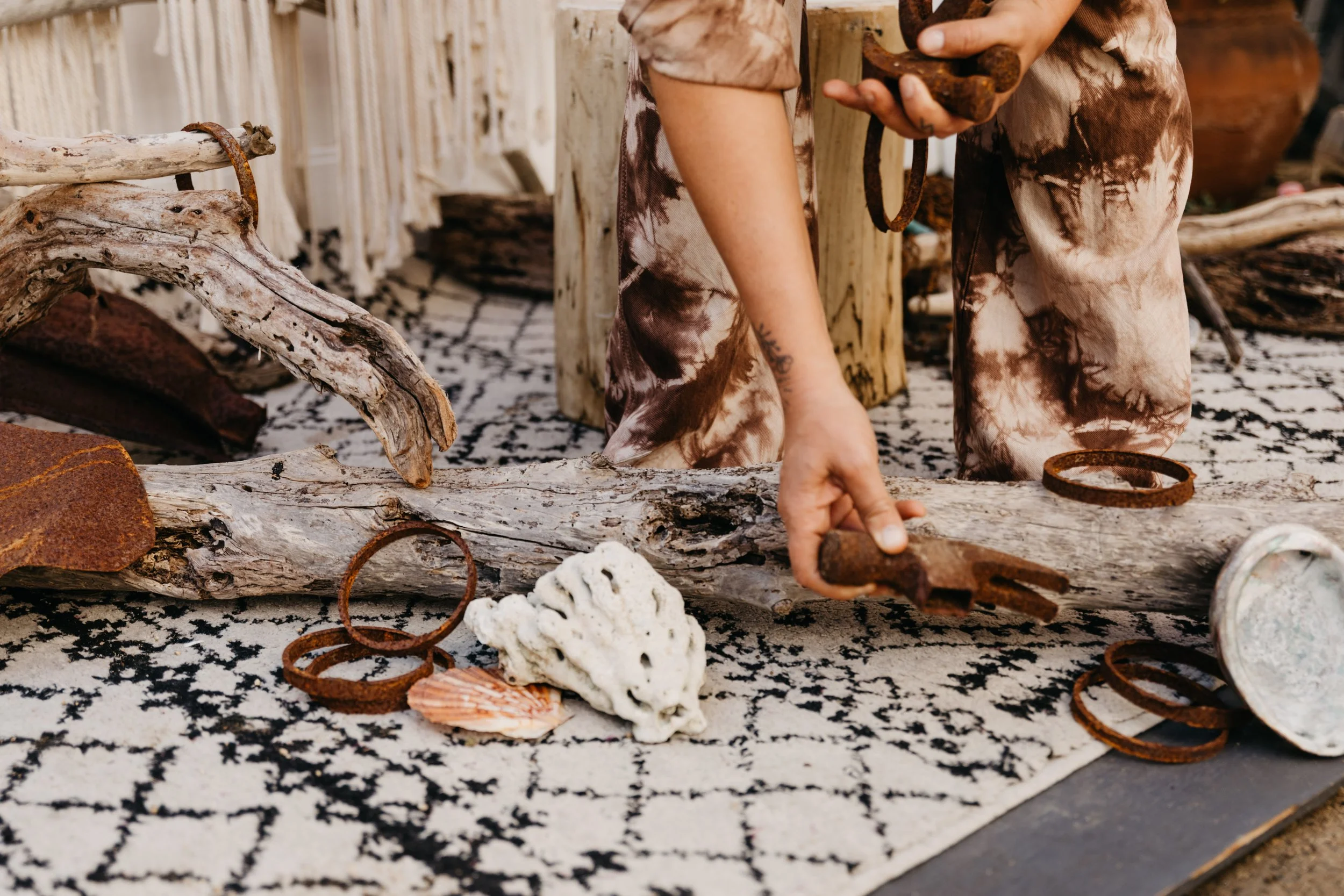 Person arranging driftwood and rusted metal rings on a patterned rug, with a terracotta pot in the background.