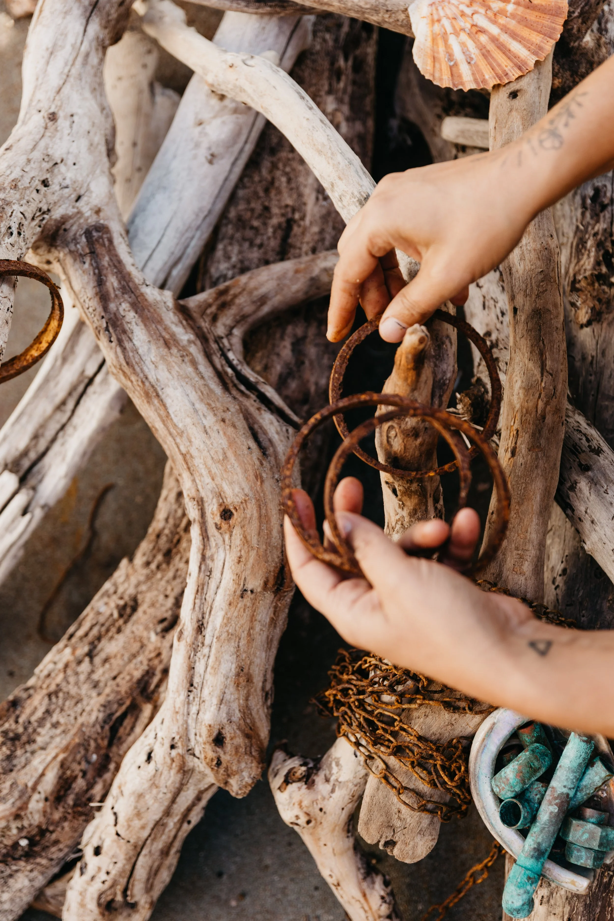 Person handling rusty metal rings on weathered driftwood with seashells and chains outdoors.