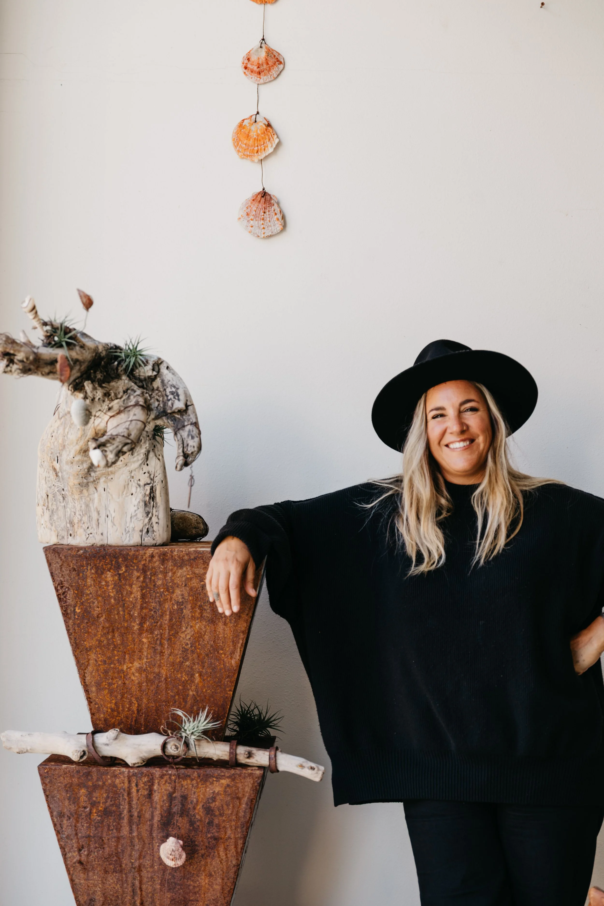 A woman with blonde hair wearing a black wide-brimmed hat and black sweater, smiling, standing next to a decorative plant arrangement on a tall, rust-colored metal stand against a light-colored wall.