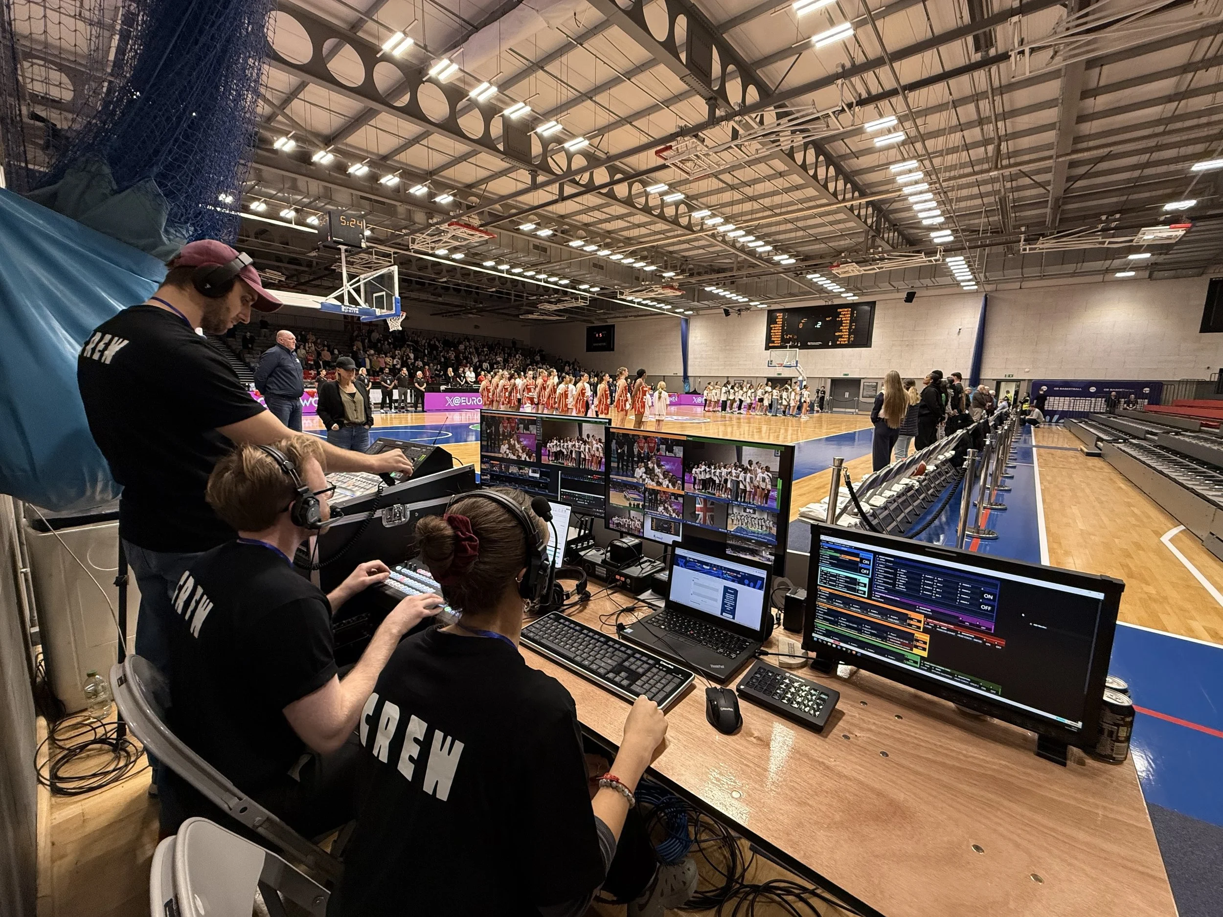 Video crew operating equipment during a basketball game in an indoor gymnasium with players lined up on the court.