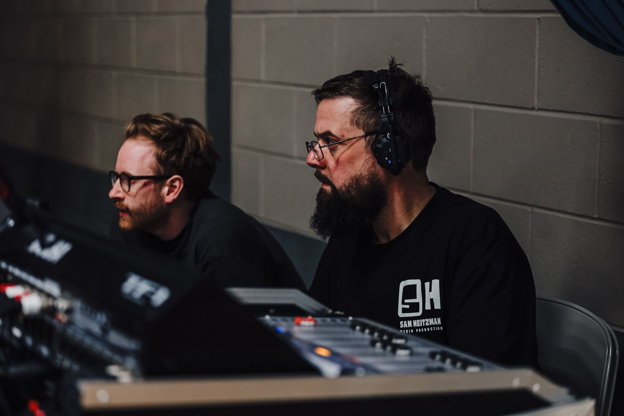 Two men sitting at a soundboard, both wearing black T-shirts and headphones. The man on the right has glasses, a beard, and short dark hair, while the man on the left has glasses, a beard, and light brown hair. They are focus on their work in a dimly lit room with a gray brick wall in the background.