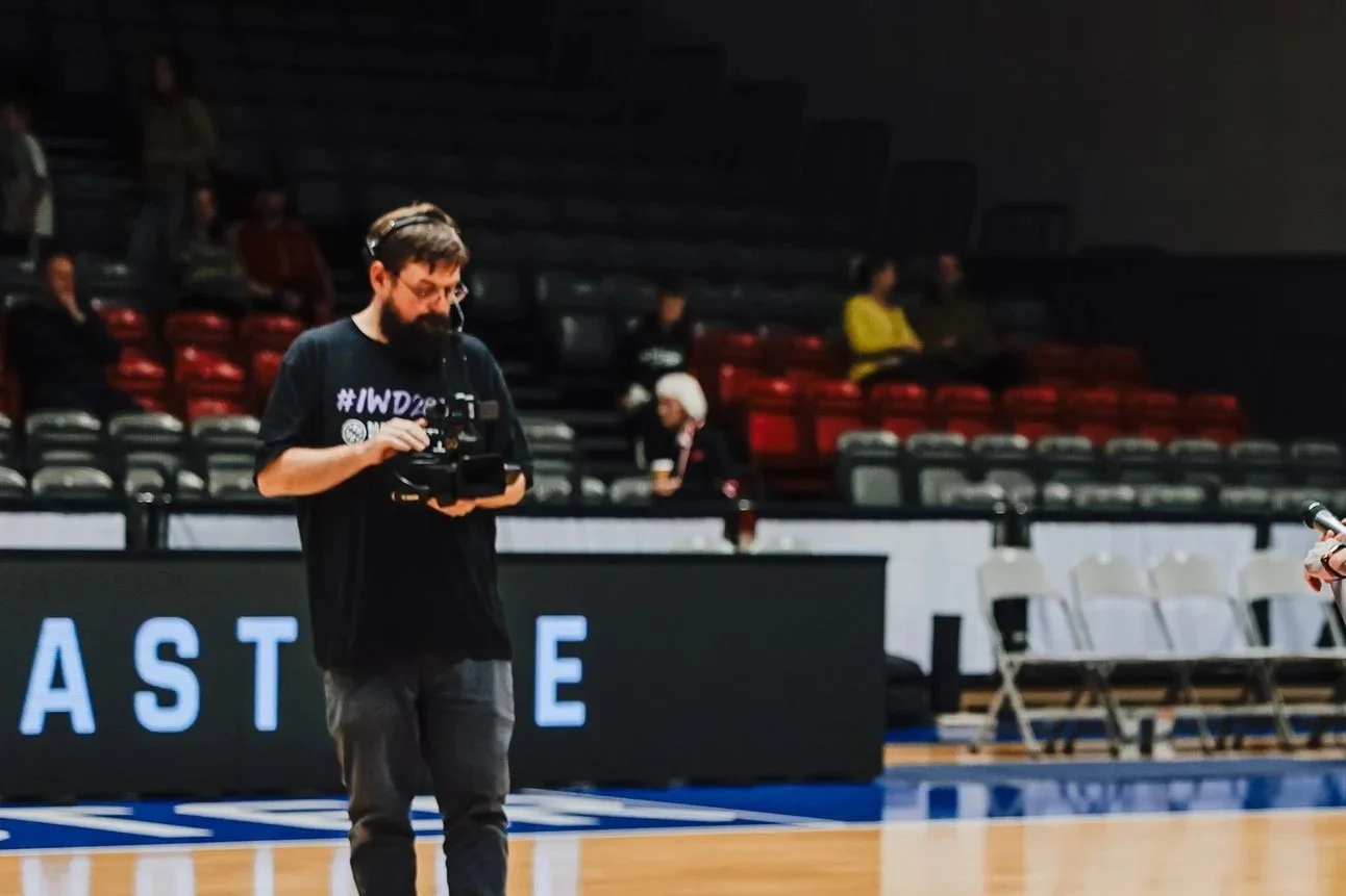 A man with a beard and glasses is operating a camera on a basketball court, with several empty chairs and a few people sitting in the background.