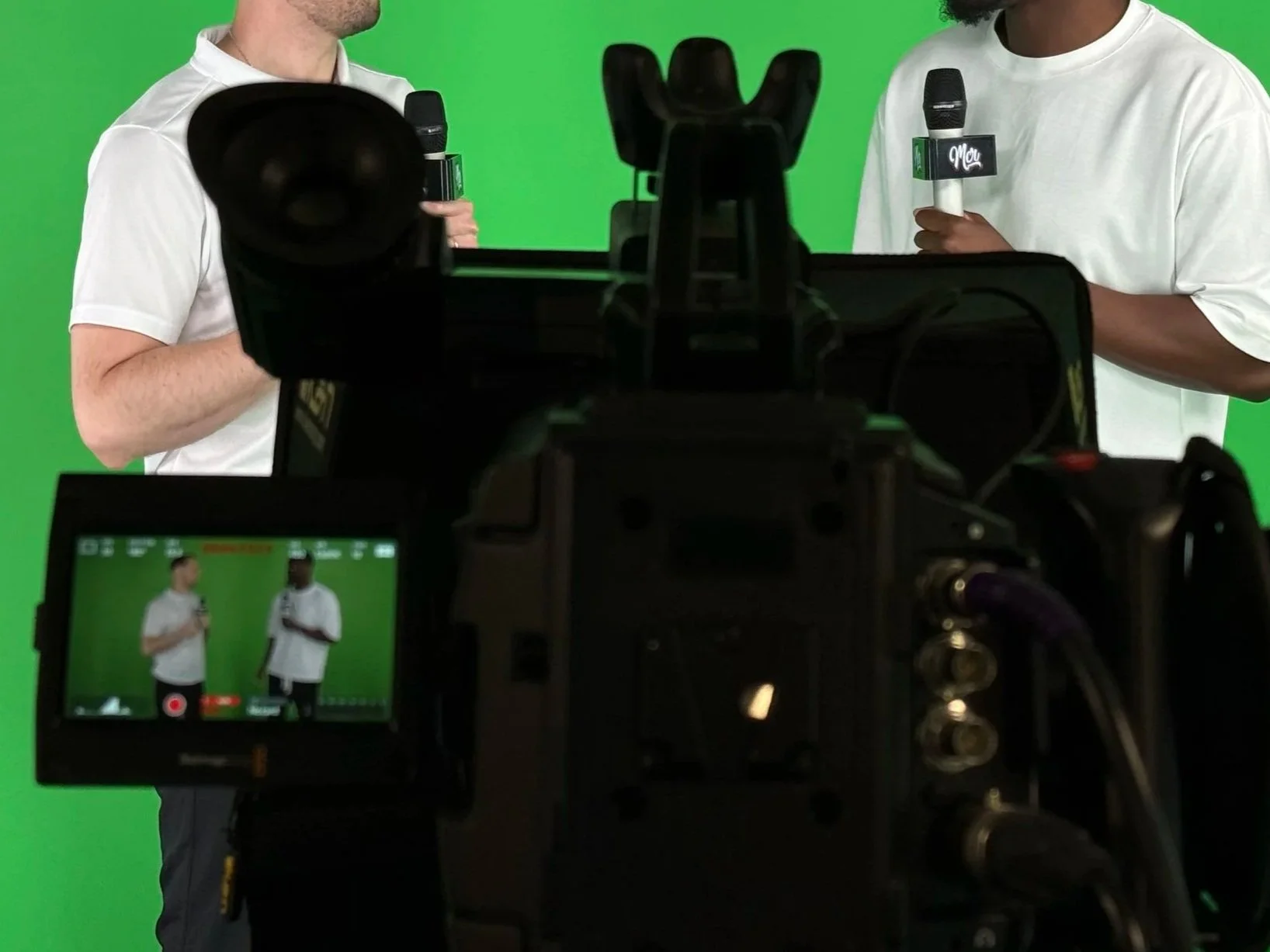 Two men are standing in front of a green screen, each holding a microphone with a label on it. A camera is recording their conversation.