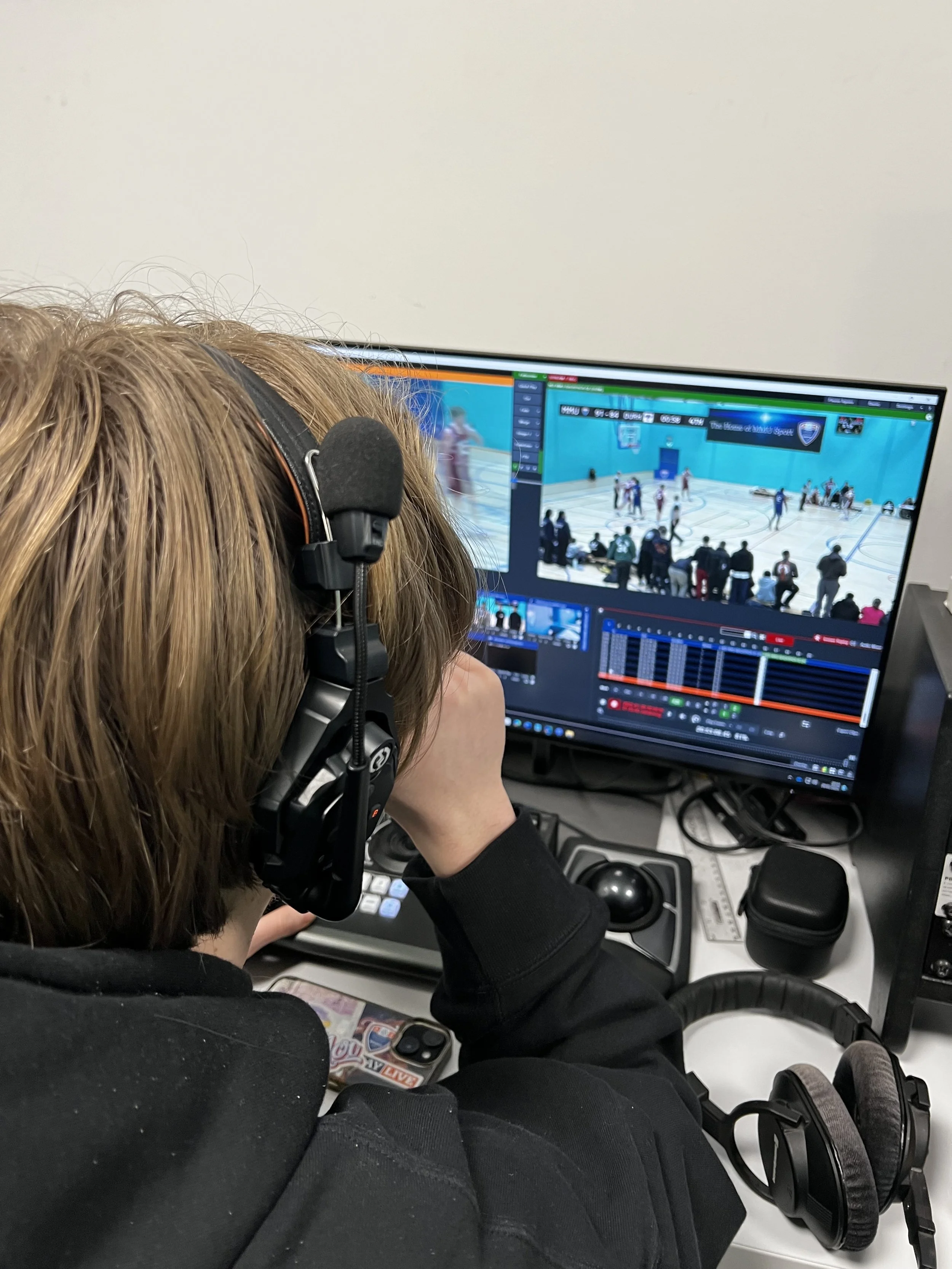 Person wearing headphones sitting in front of a computer monitor showing sports game footage, with editing software interface visible on the screen.