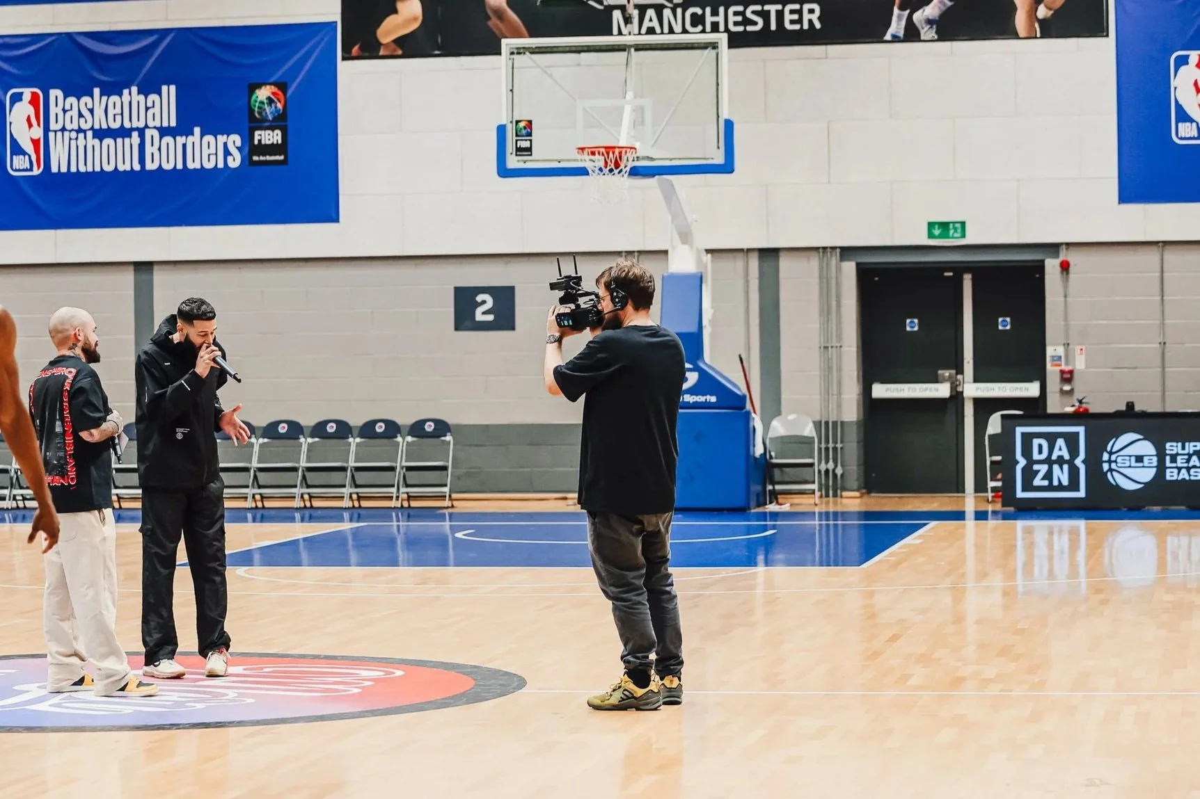 A person filming a Interview on basketball court with two men, one holding a microphone, other standing, and a camera operator recording the scene. The court has banners, including one that says "Basketball Without Borders" and another for "DAZN" and "Super League Basketball."