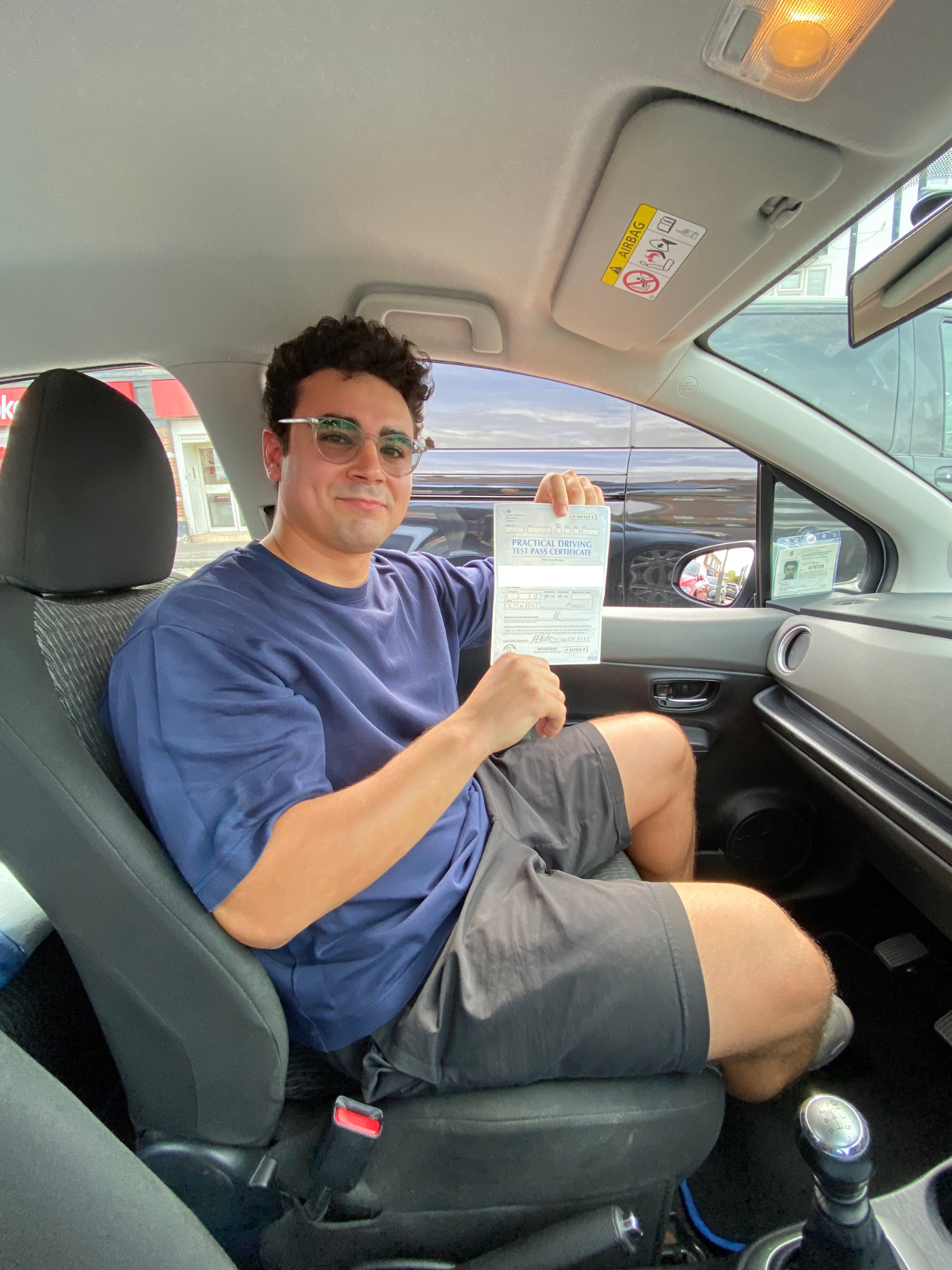 Young man sitting in car driver seat holding up a practical driving test pass certificate.
