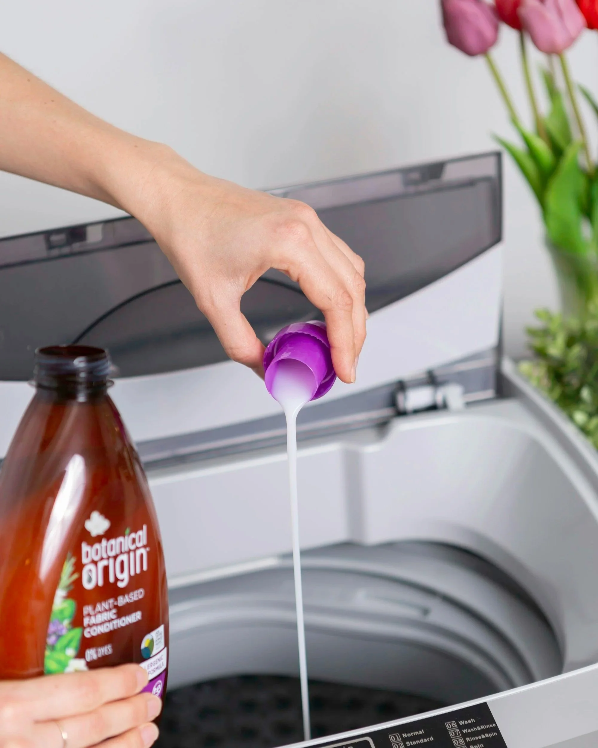Person pouring liquid fabric conditioner from purple bottle into washing machine