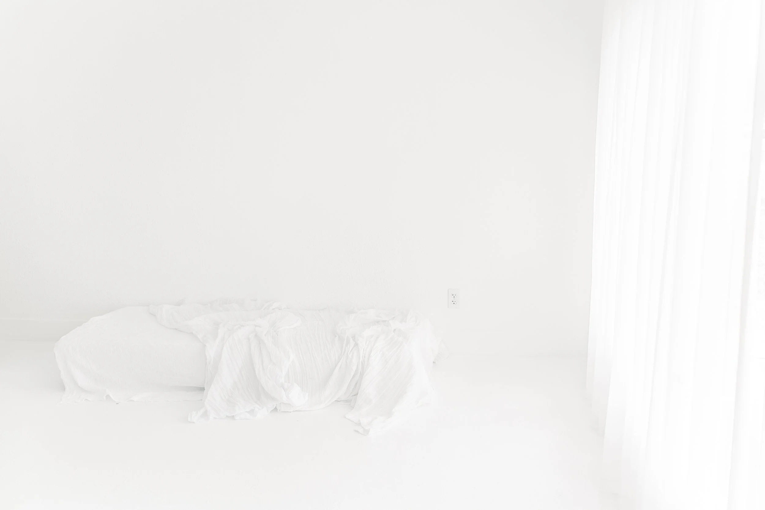 A minimalistic white room with a low bed covered in white sheets and a sheer white curtain on the right side.