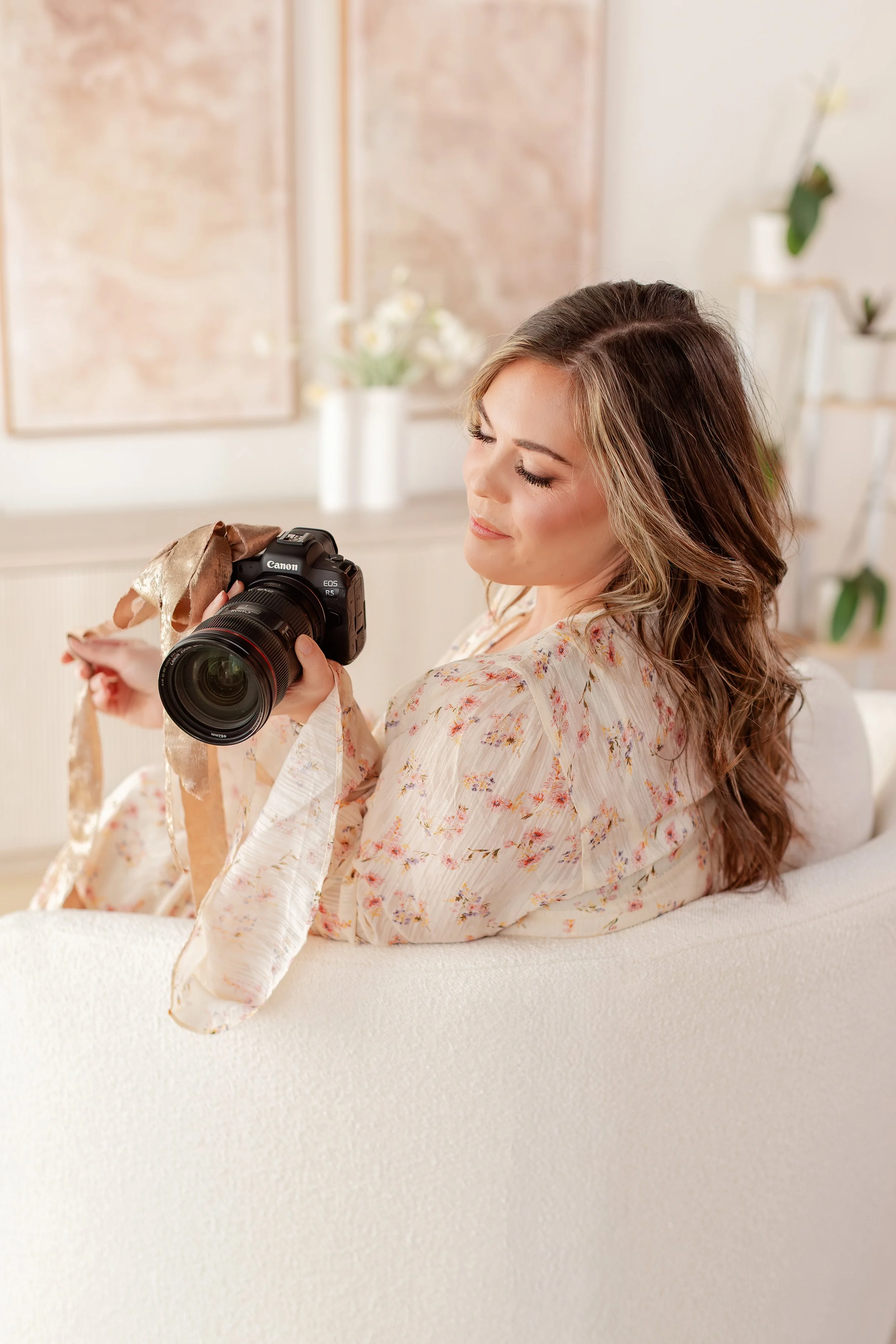 A woman with long, wavy brown hair is sitting on a white sofa, looking at a Canon camera she is holding in her hands, inside a light-colored room with floral decor.