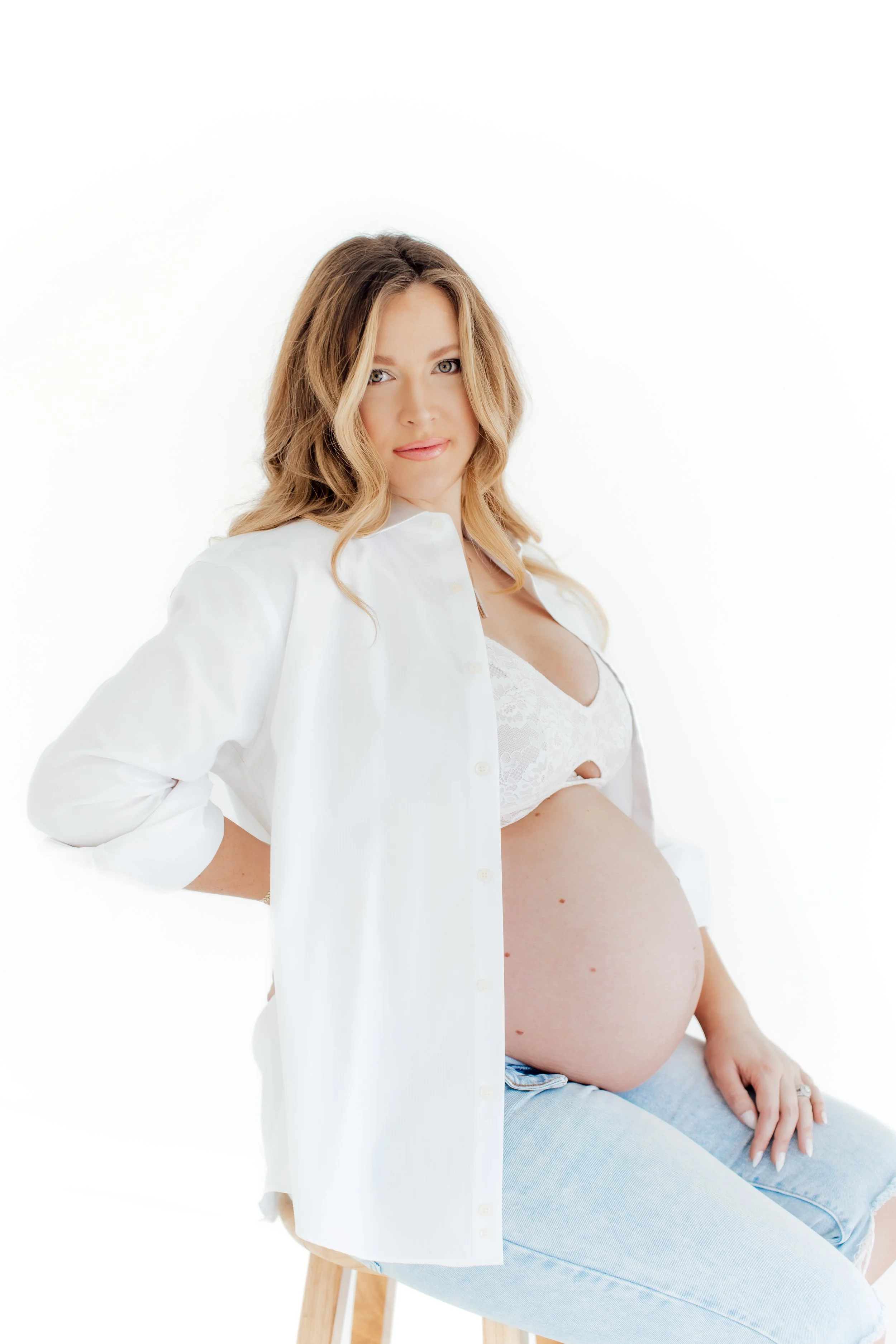 Pregnant woman sitting on a stool wearing a white open shirt and lace bra, with a neutral expression, against a plain white background.