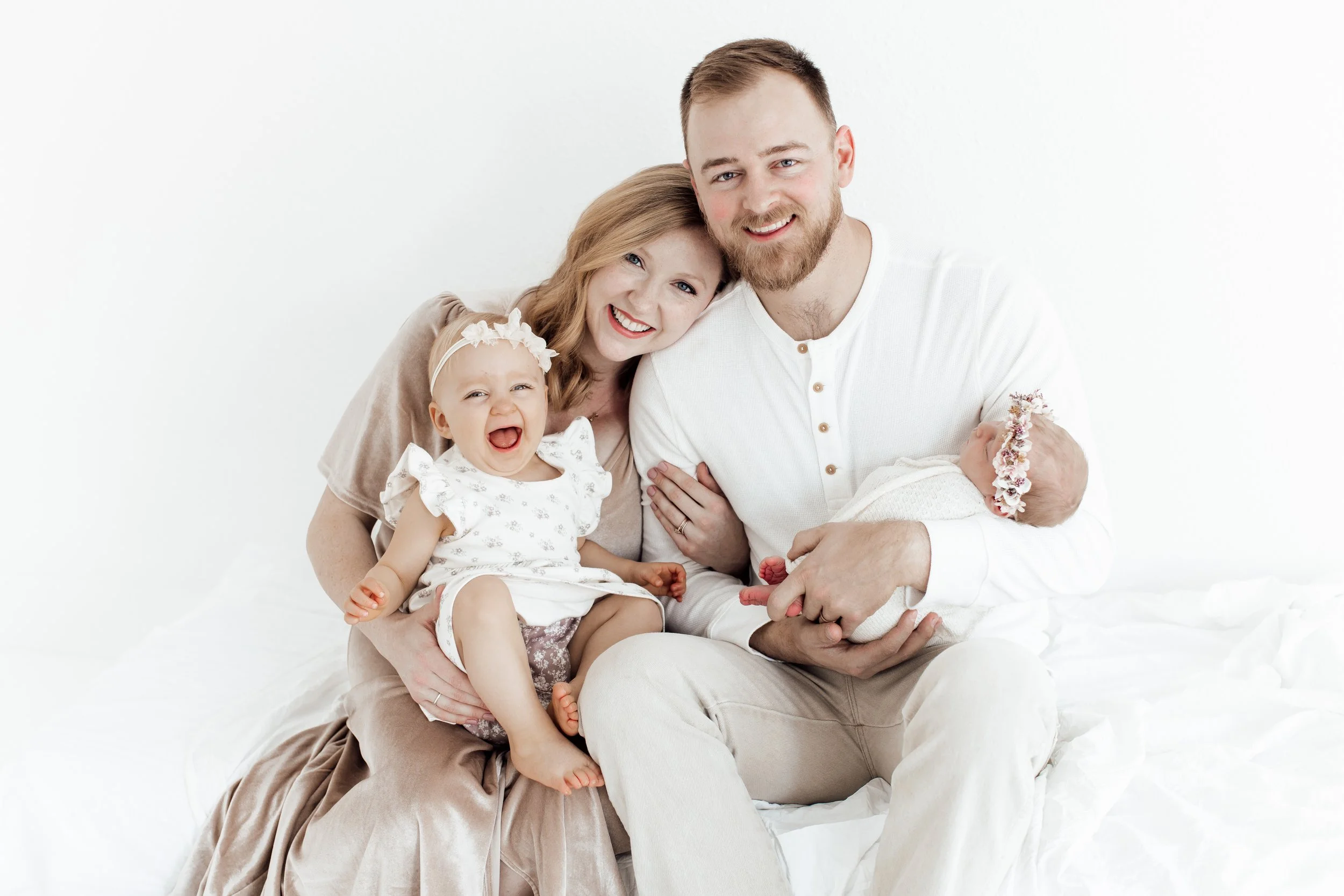 Family portrait of a happy couple with two young children, one girl and a newborn, sitting on a white bed in a bright room.