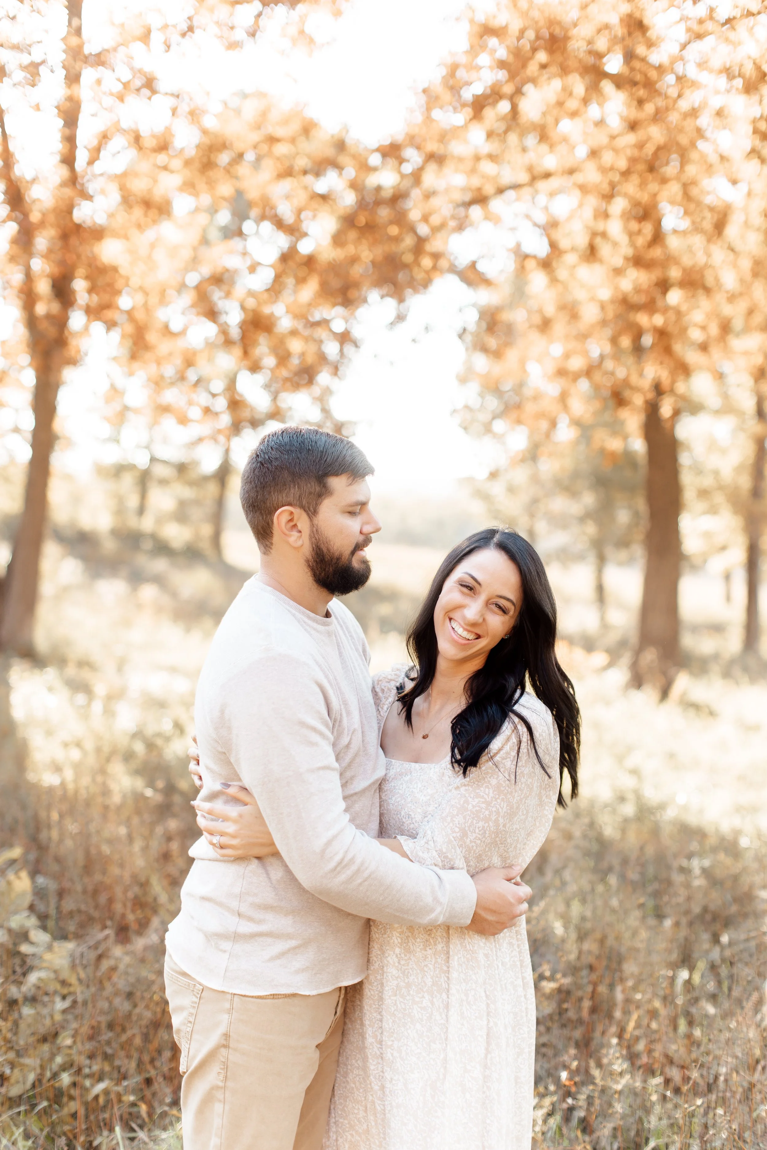 A happy couple standing outside in an autumn setting, with trees and warm sunlight in the background.