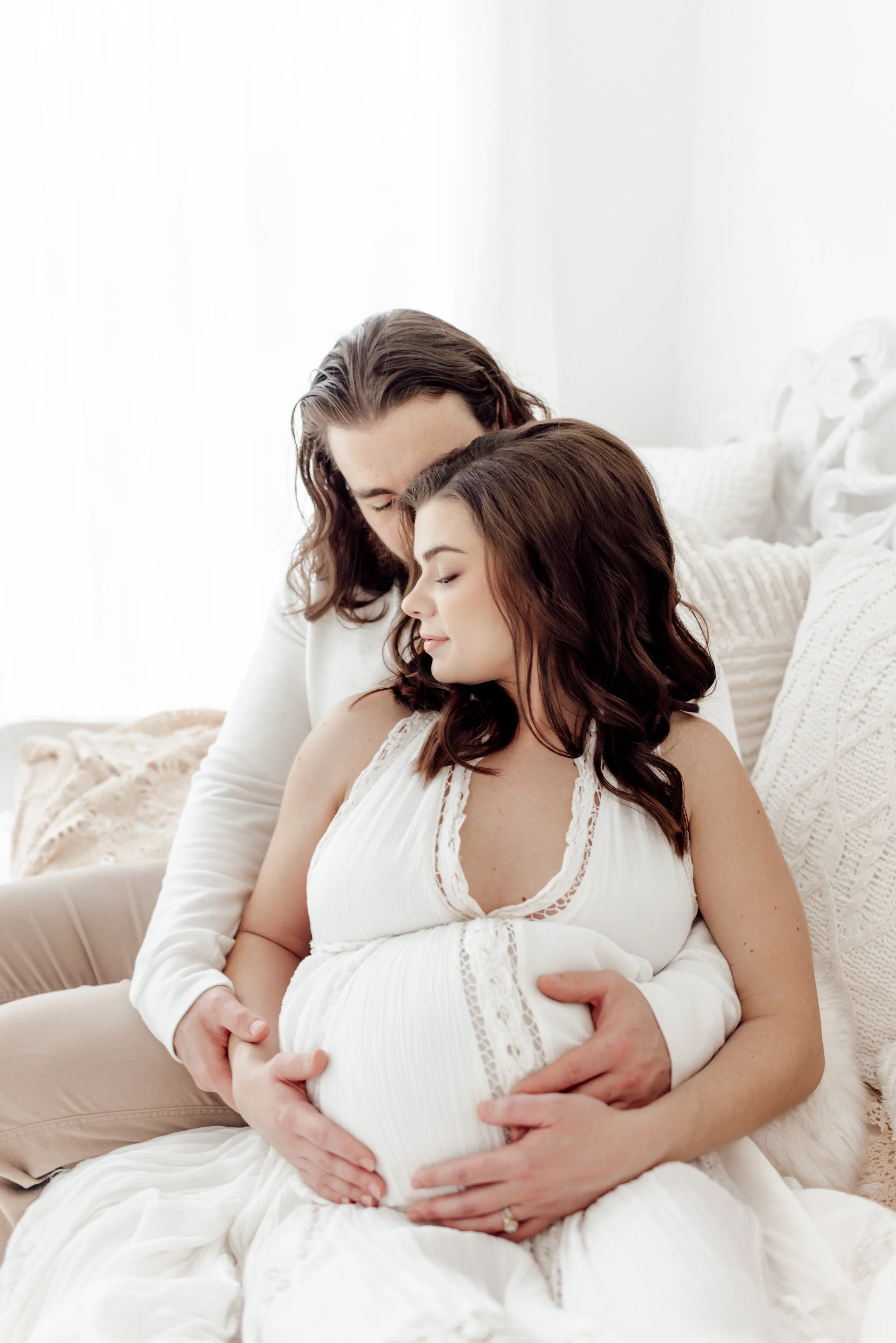 A couple sitting close together on a bed, with the woman being pregnant, gently cradling her belly.