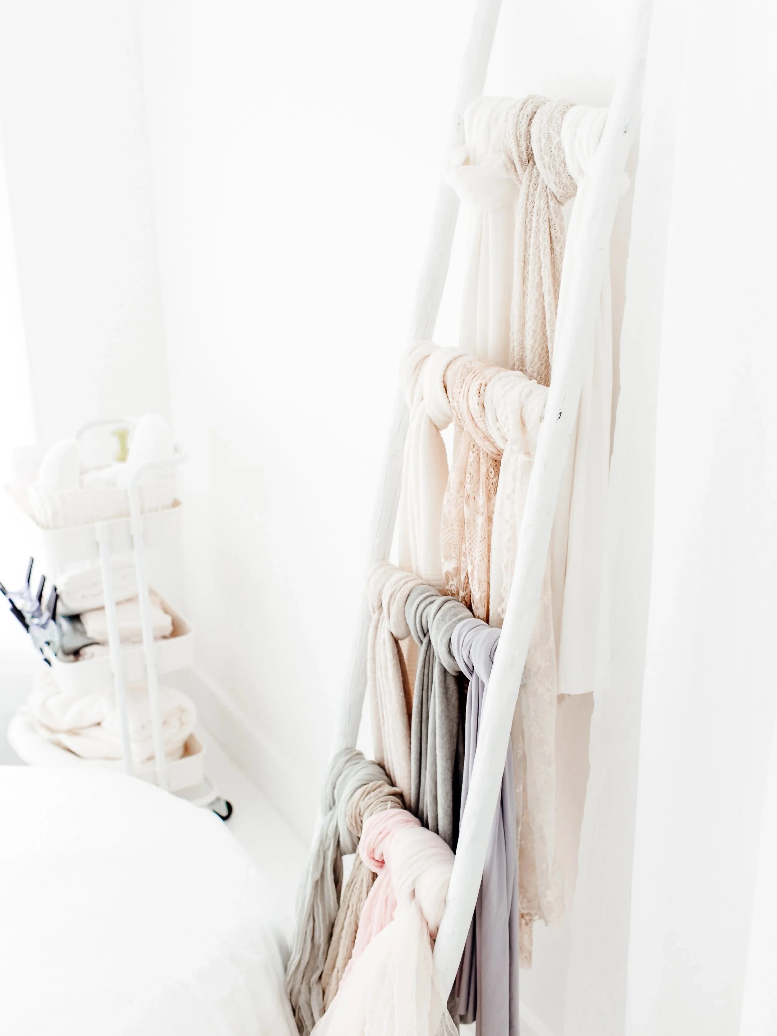 A white ladder decorated with pastel-colored scarves or fabric items hanging from its rungs, positioned in a bright room with white walls.