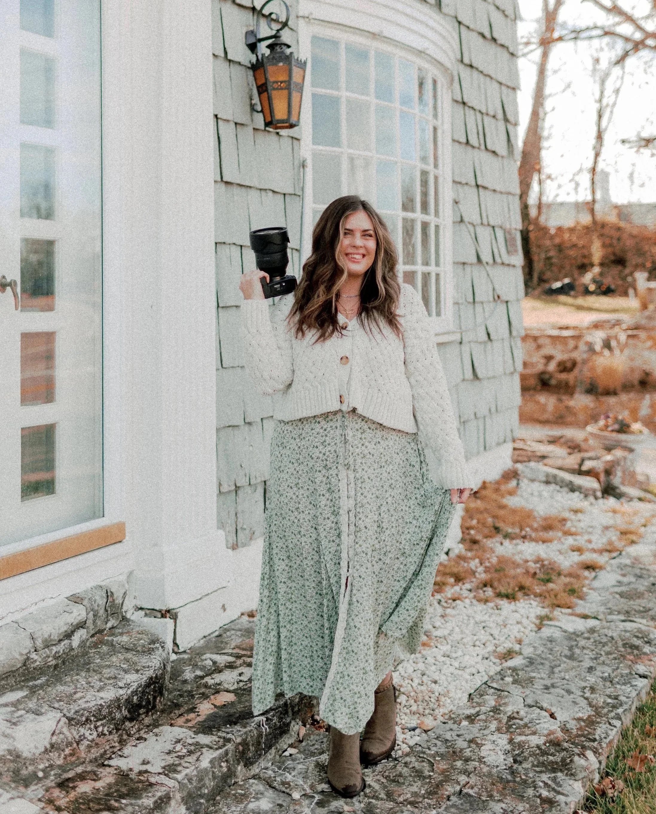 A young woman with long, wavy brown hair, smiling, holding a camera with a large lens. She is standing outside next to a stone path and a house with grey shingle siding. She wears a cream-colored knit sweater, patterned wide-leg pants, and brown ankle boots. The background features autumn trees and a yard.