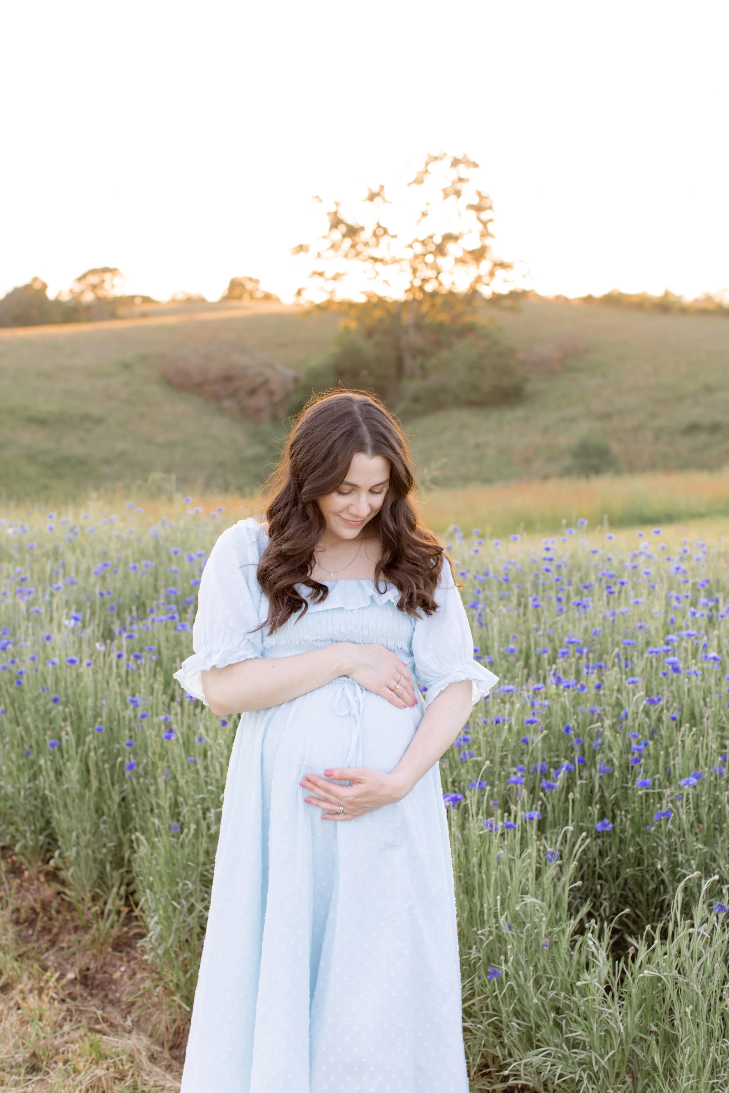 A pregnant woman standing in a field of purple flowers during sunset, gently holding her belly and smiling.