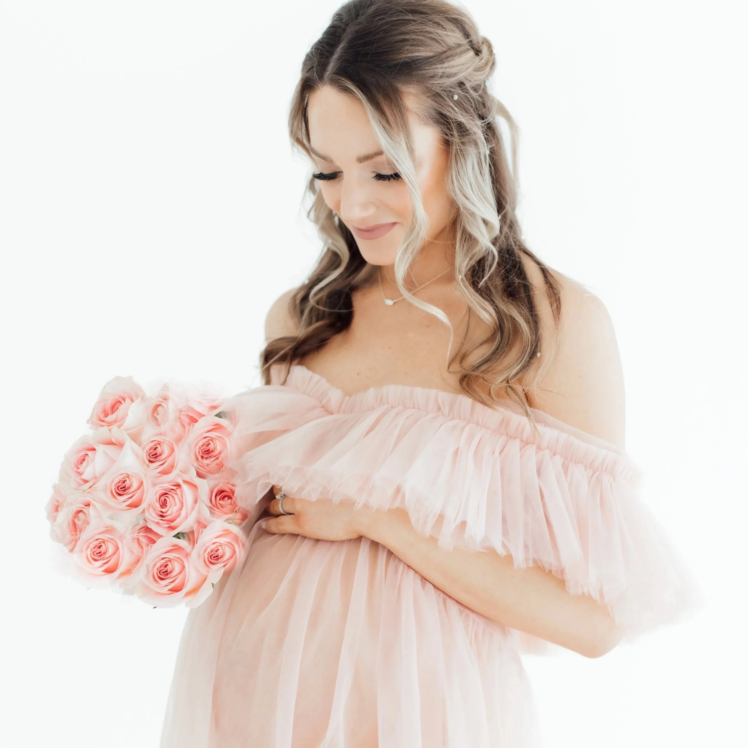 Pregnant woman in a pink off-shoulder dress holding a bouquet of pink roses, smiling softly with her eyes closed.
