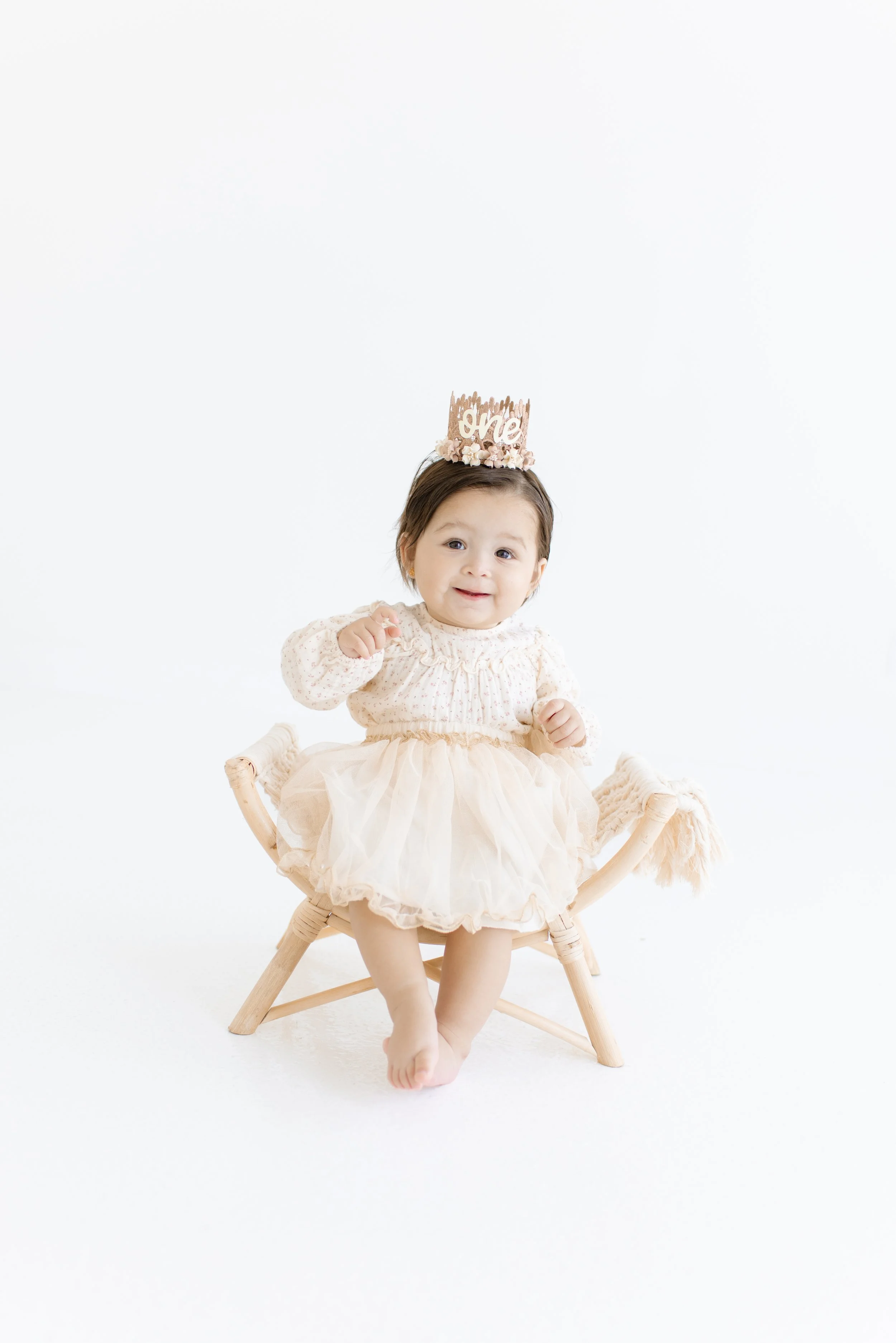 Young girl celebrating her first birthday, wearing a beige dress with a tulle skirt and a birthday crown with 'one' written on it, sitting on a small wooden chair against a white background.