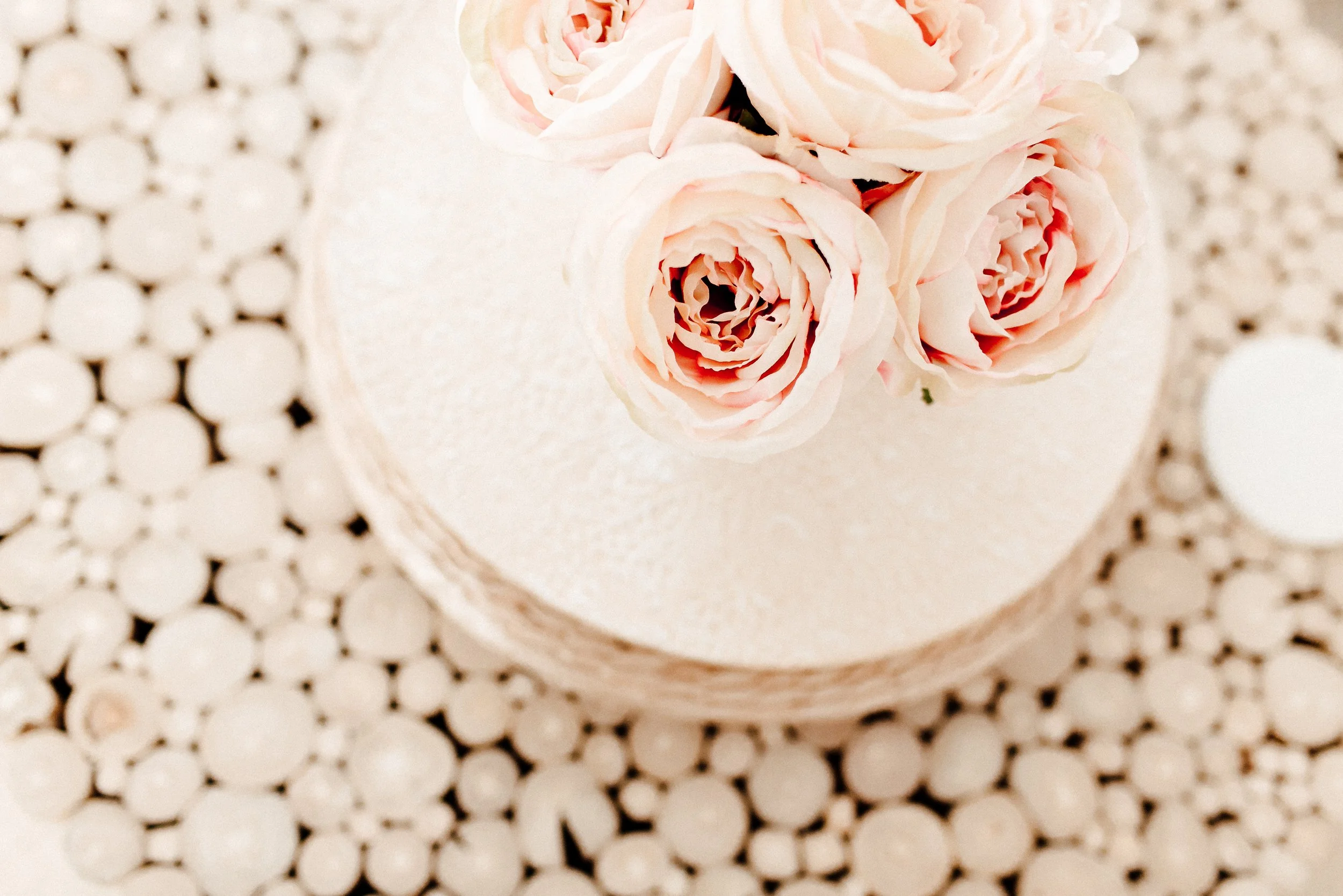A round cake decorated with a bouquet of light pink roses on top, placed on a table covered with a pattern of small white circular objects.