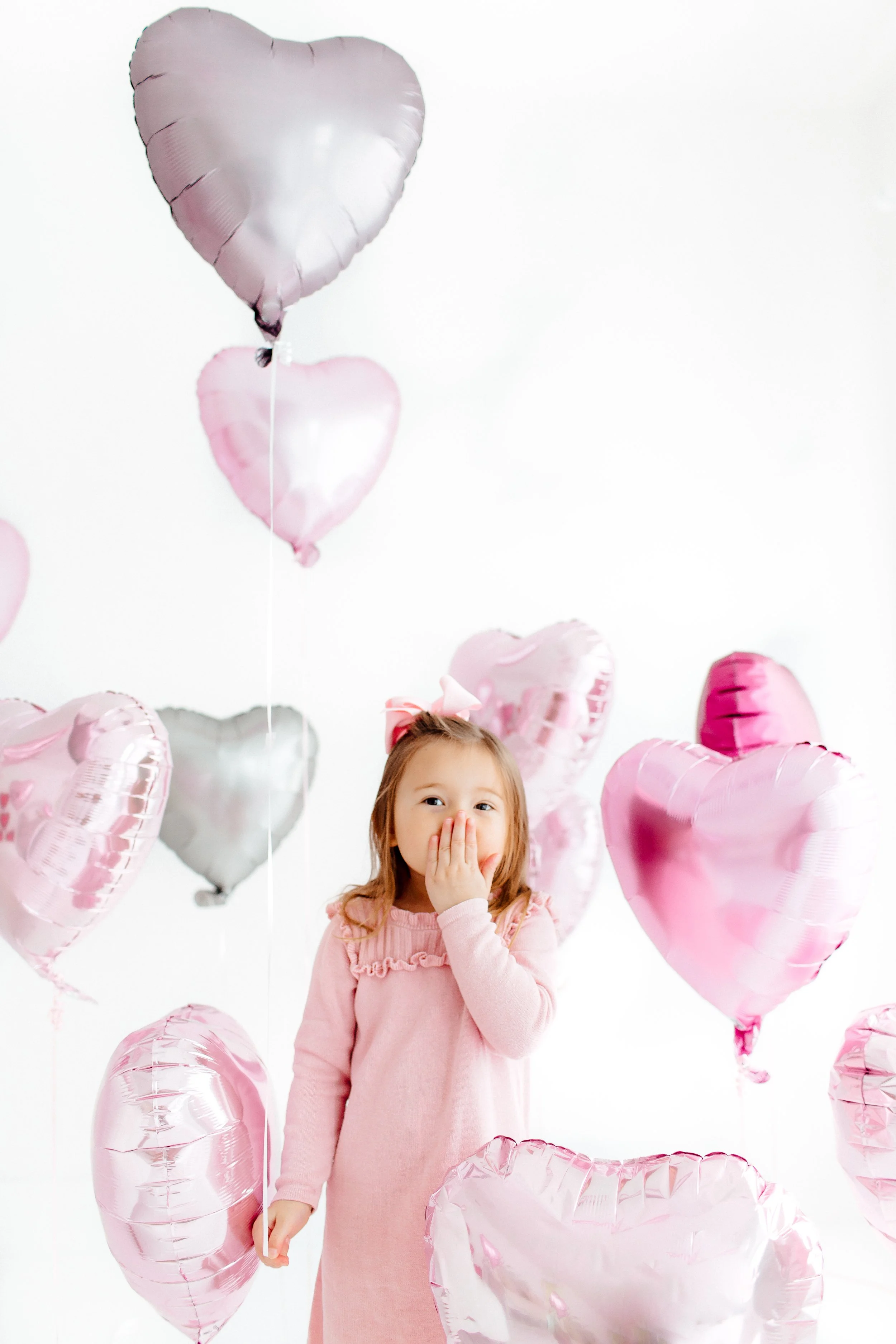 A young girl with a pink dress and bow, surrounded by pink and silver heart-shaped balloons, is covering her mouth with her hand in a playful gesture.