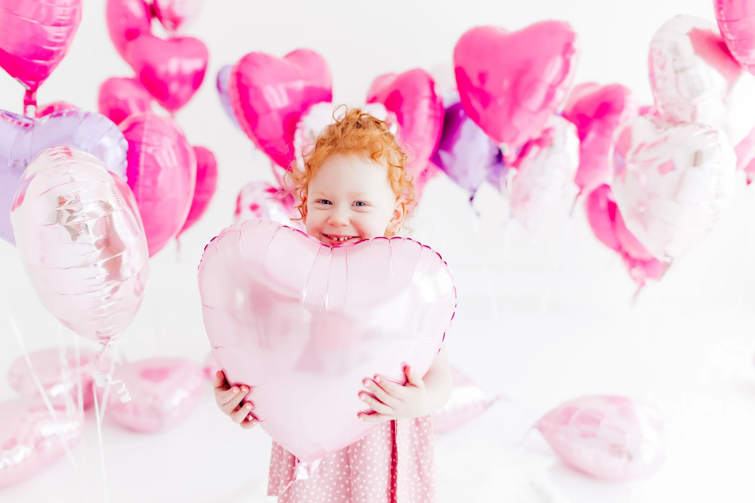 A young girl with curly red hair holding a large pink heart-shaped balloon, surrounded by pink and white heart-shaped balloons, on a white background.