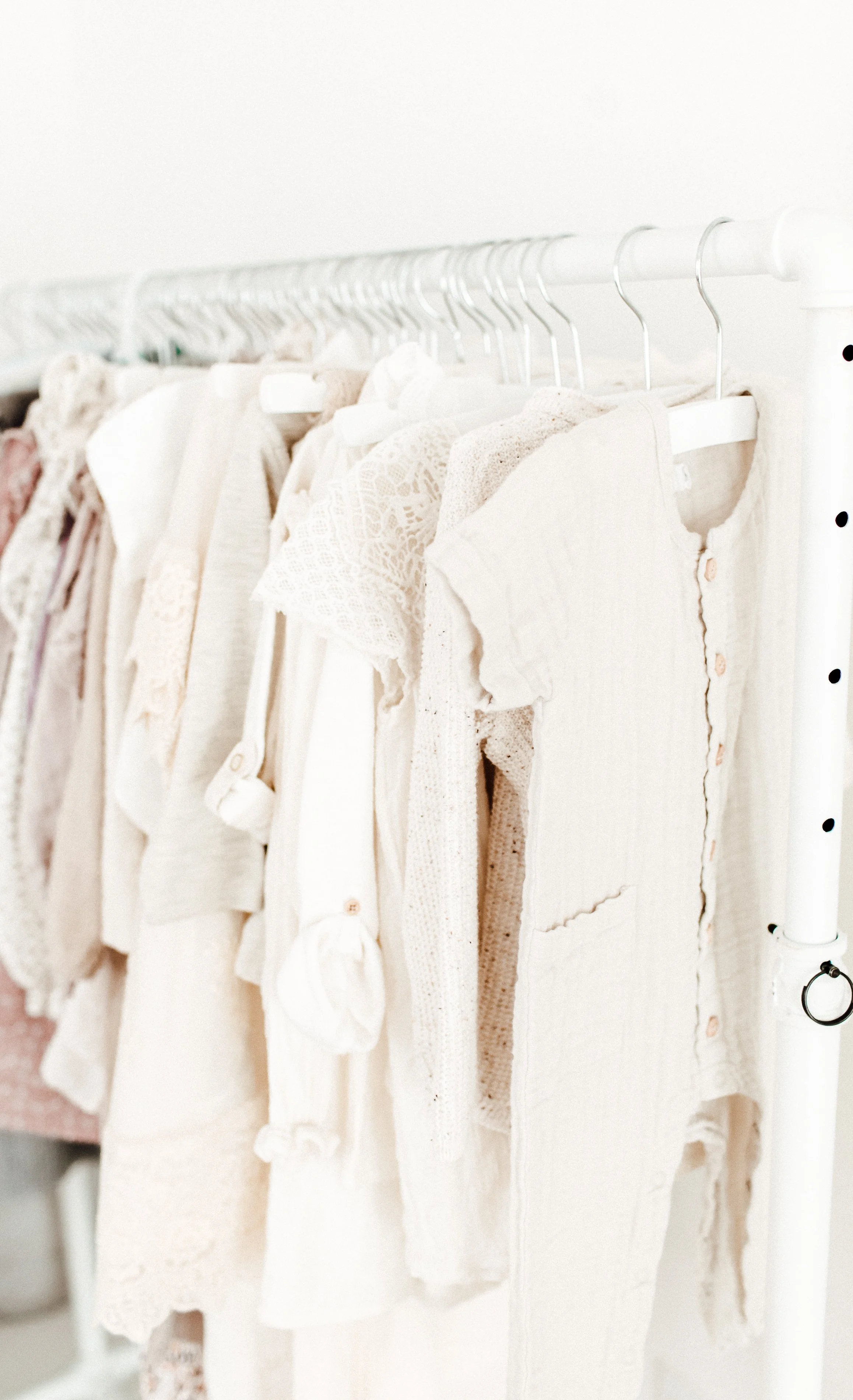 Clothes hanging on a white rack, mostly in light, neutral colors like white, beige, and cream, with some lace and crochet textures.