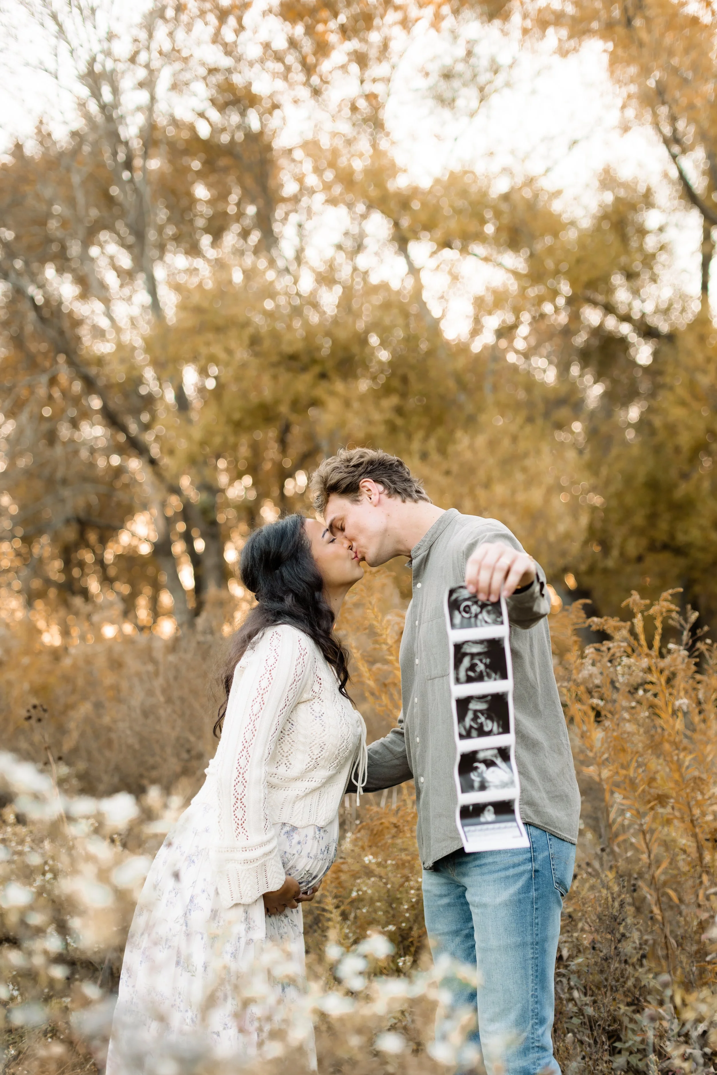 A couple sharing a kiss outdoors during autumn, with the man holding ultrasound images, surrounded by trees with orange and yellow leaves.