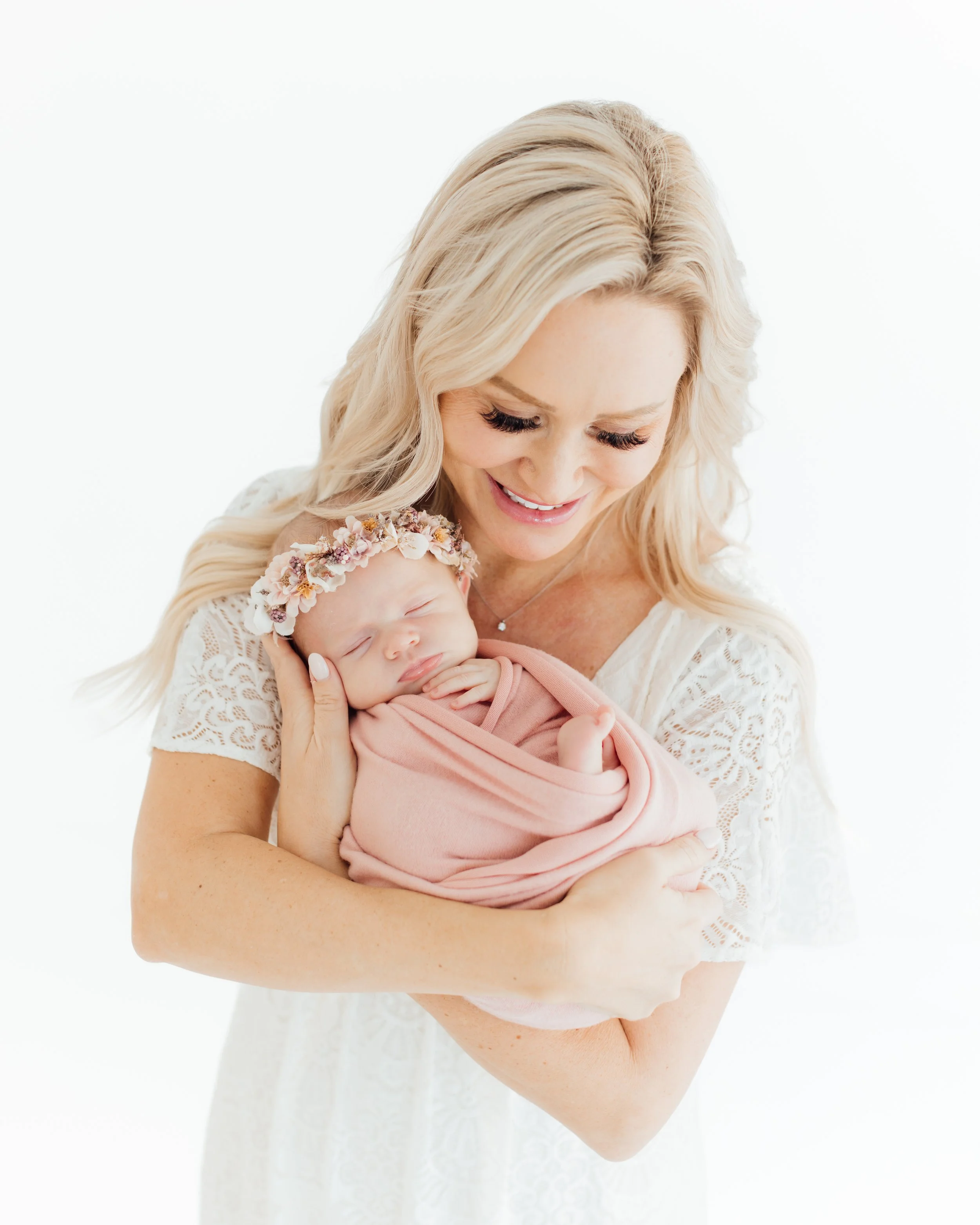 A woman with long blonde hair smiling and holding a sleeping newborn baby wrapped in pink cloth, wearing a floral headband, against a white background.