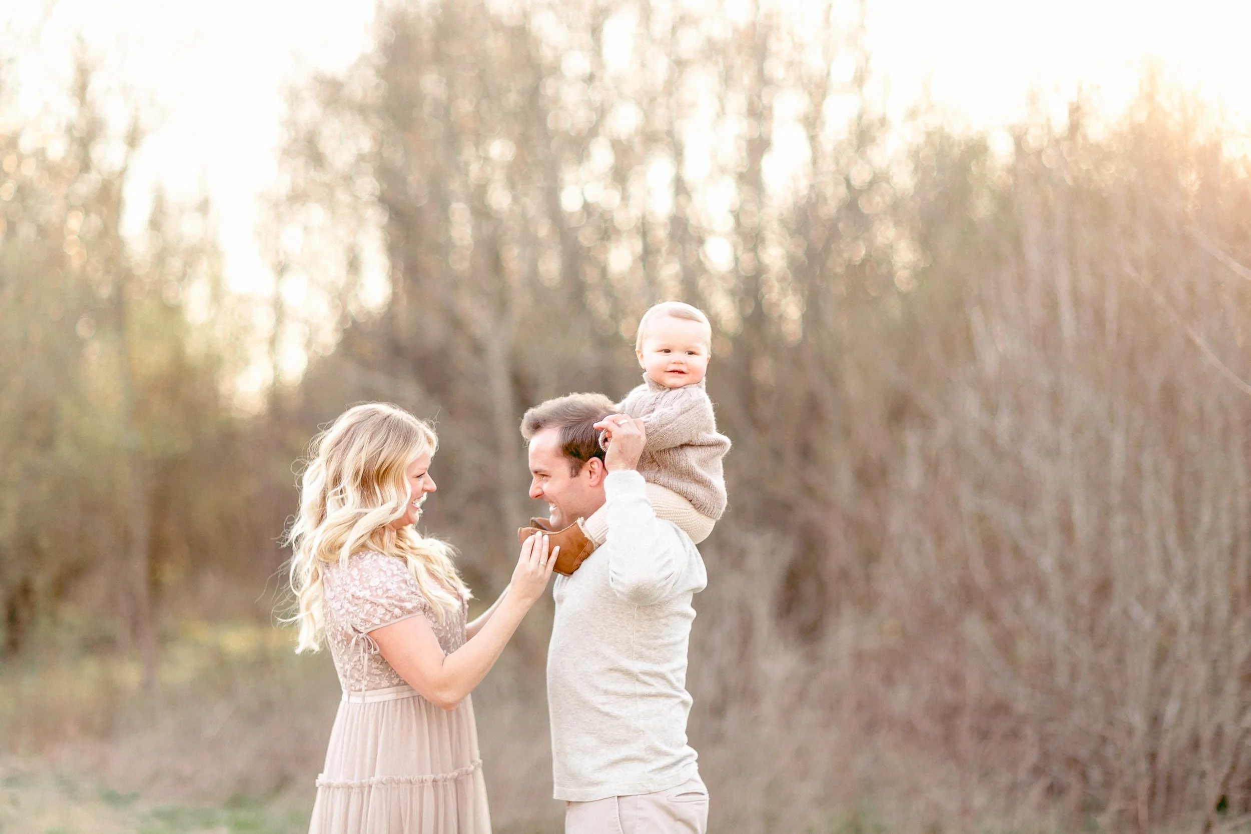 A family of three outdoors in a wooded area during sunset, with the father carrying a baby on his shoulders and the mother holding a snack near the baby's mouth, all smiling and sharing a joyful moment.