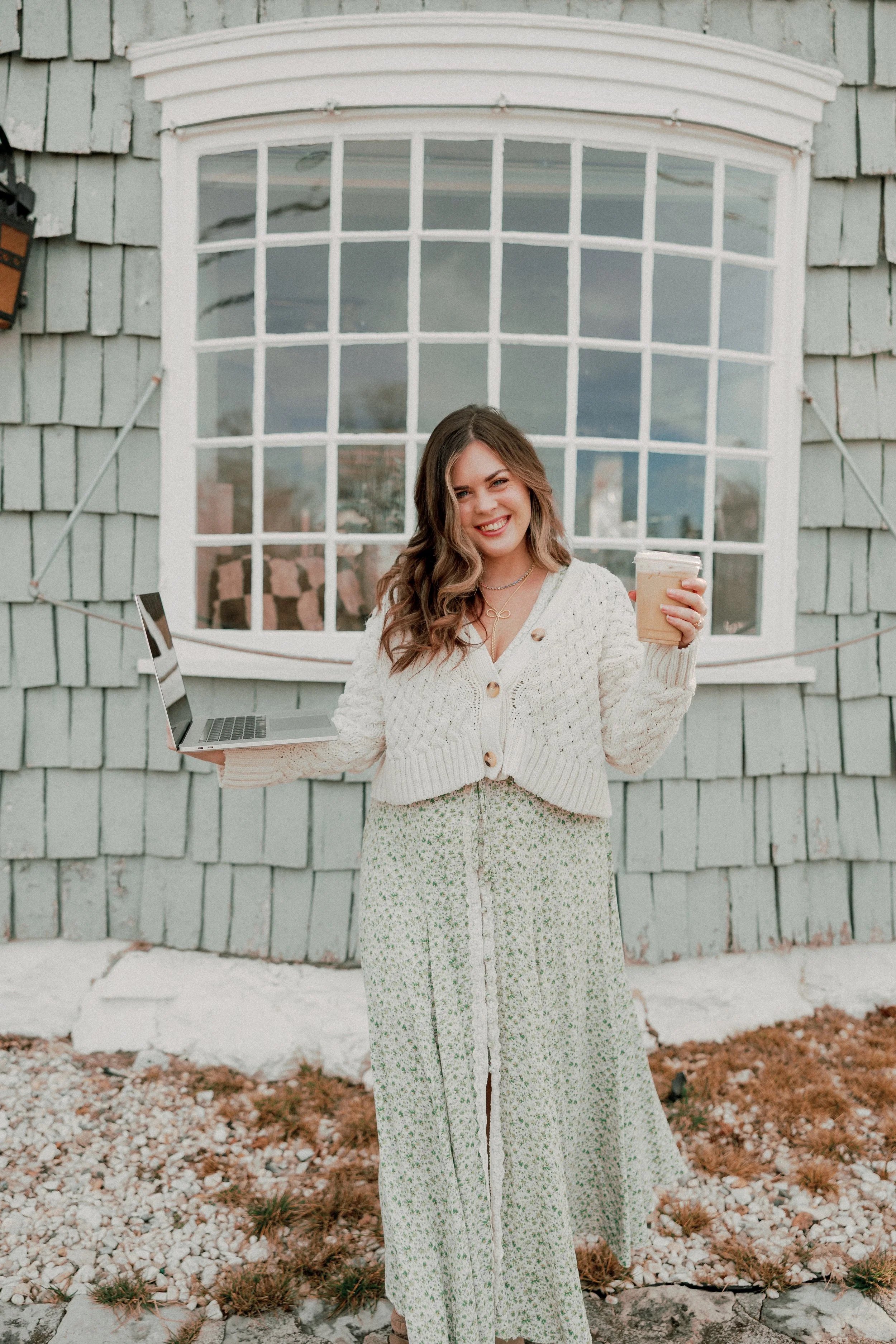 A young woman smiling and holding a laptop in one hand and a iced coffee in the other, standing outdoors in front of a house with green shingle siding and a large window.