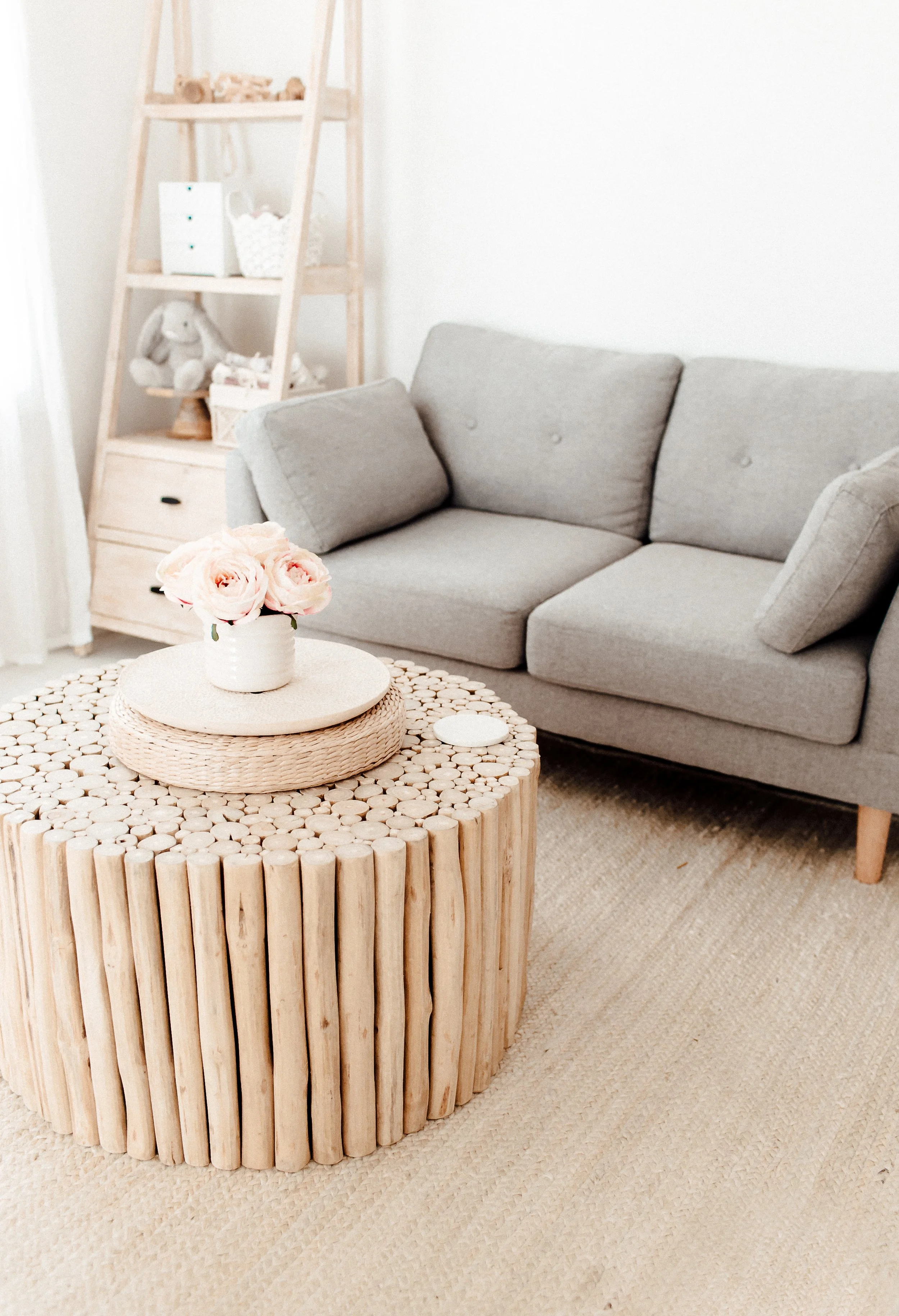 A cozy living room with a gray fabric sofa, a round wooden coffee table with a basket weave design, and a wooden ladder-style shelf with some toys and decor in the background. The coffee table has a white vase with light pink roses.