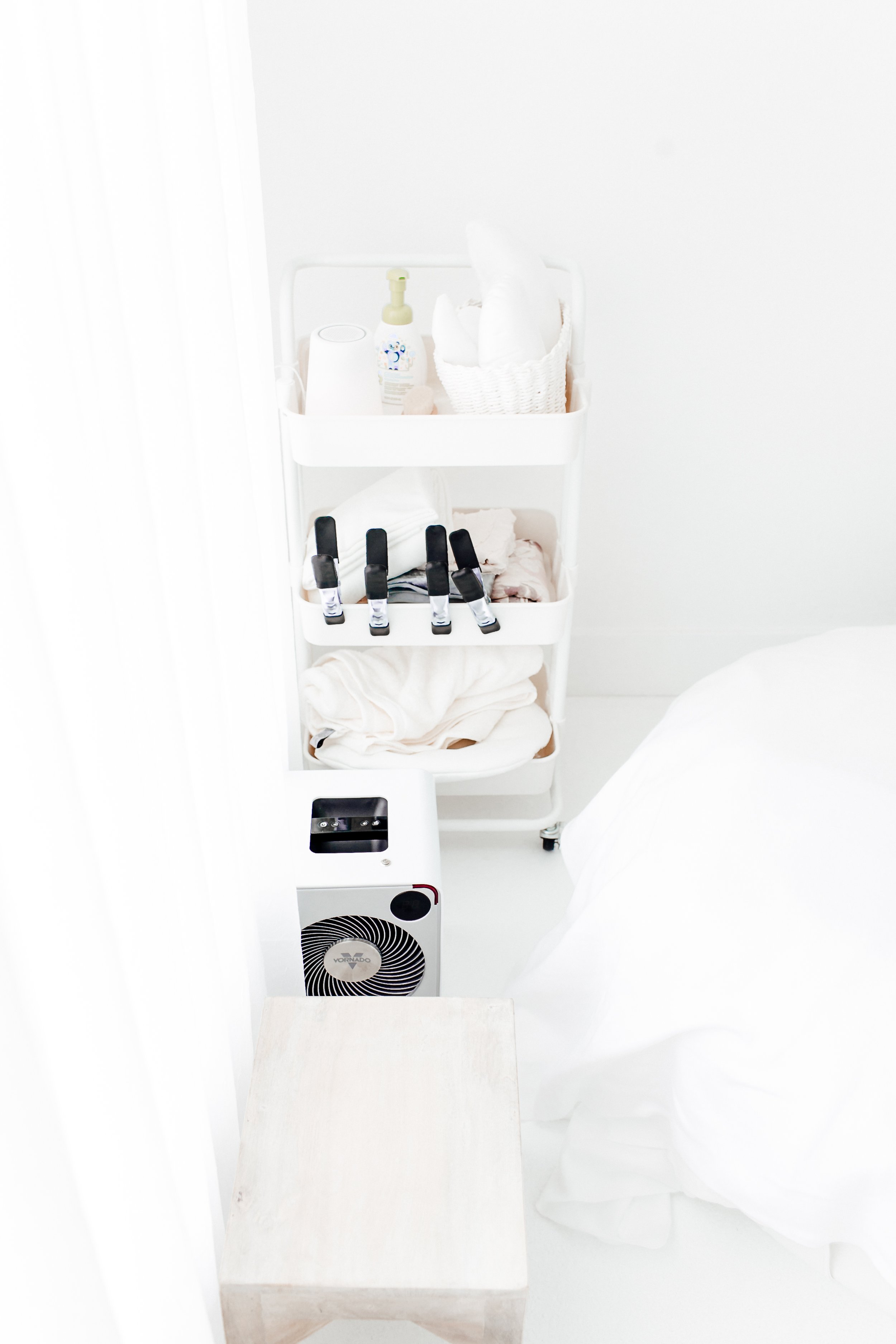 A white rolling cart with three shelves holding personal care items and clothes, next to a bed with white bedding, a small white table in front, and a white air purifier on the floor.