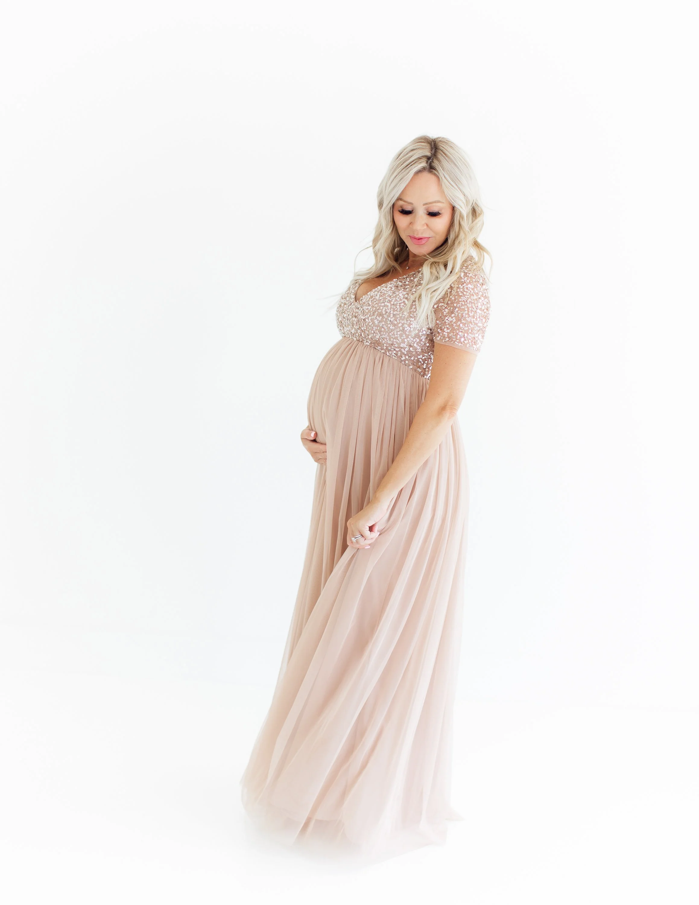 Pregnant woman in a long, blush pink dress with sequin top, looking down and holding her belly, standing against a plain white background.
