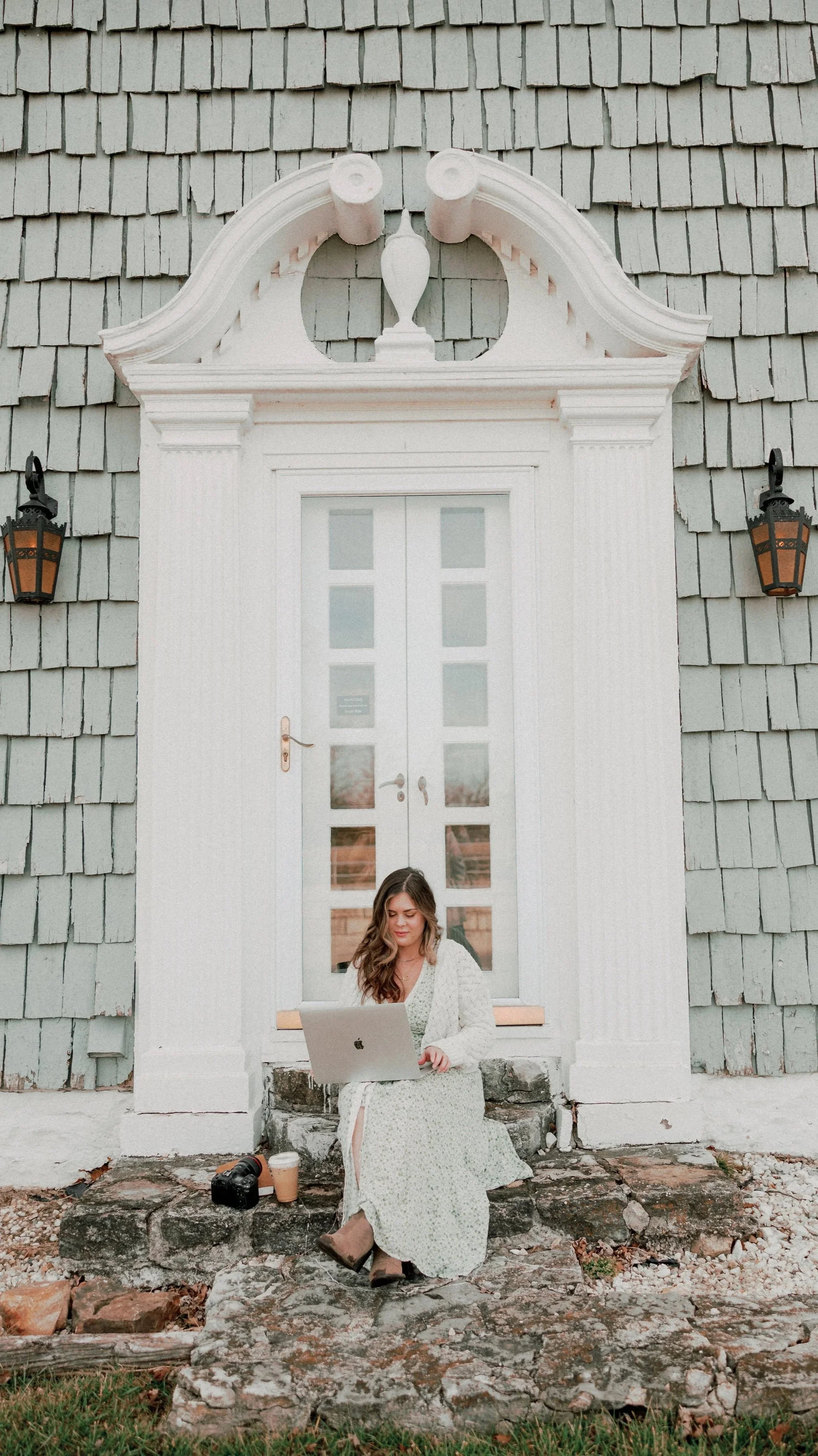 A woman sitting on a stone step in front of a white door with glass panes, using a laptop, with a camera and drink beside her, outside a house with gray shingles and black lanterns.