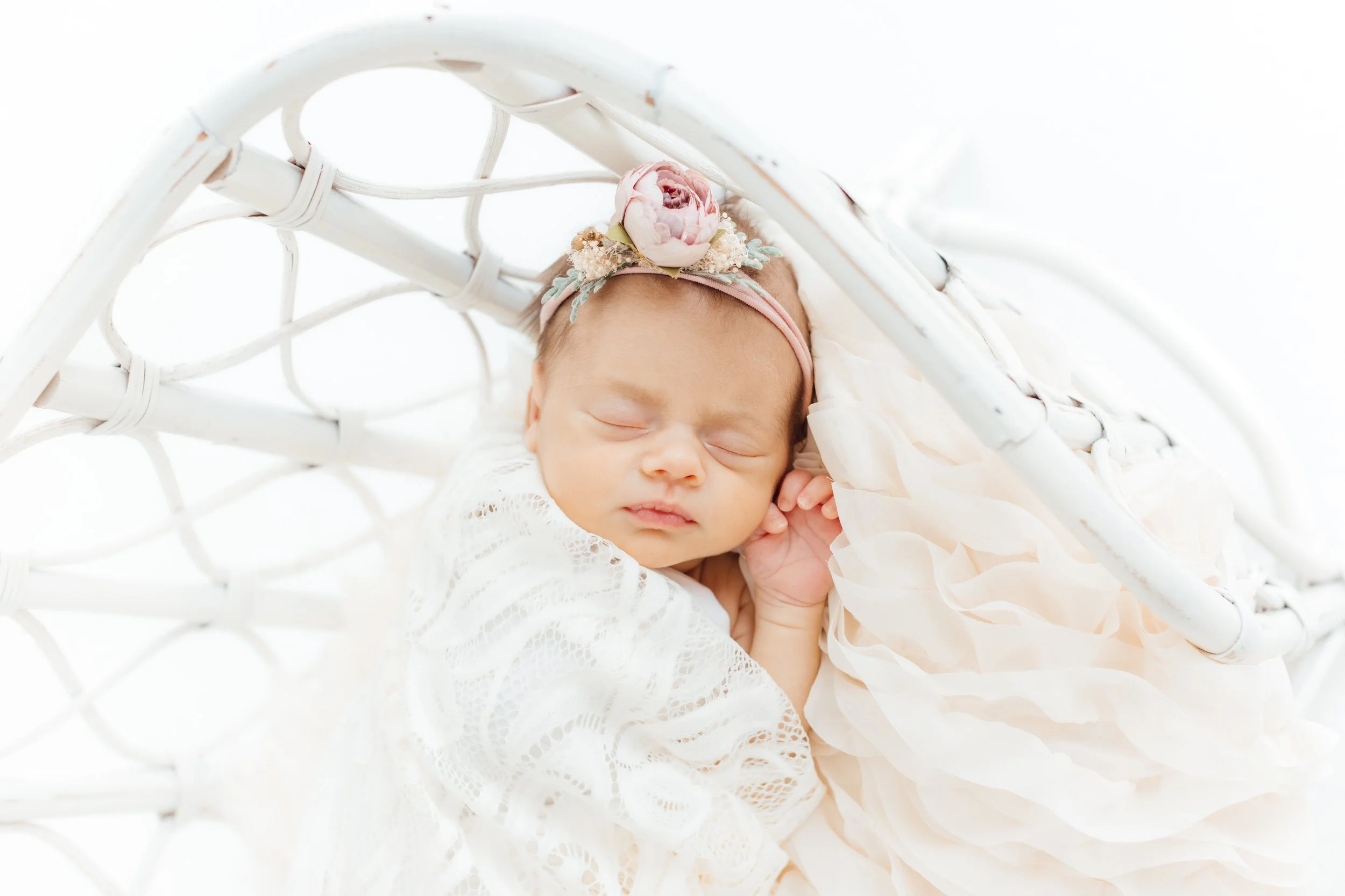 A sleeping newborn baby girl with a floral headband, lying in a white wicker basket with a soft blanket.